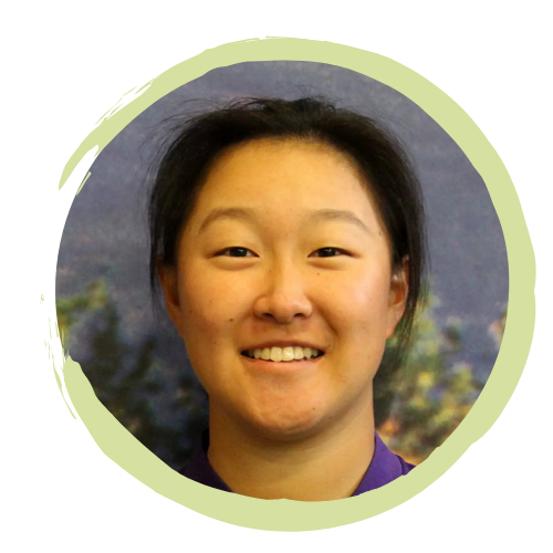 Smiling woman with short dark hair, standing outdoors with mountains and trees in the background.