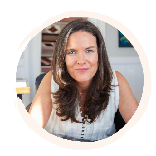 A woman with brown hair and blue eyes, smiling at the camera, wearing a sleeveless white blouse, sitting at a desk in an office setting.