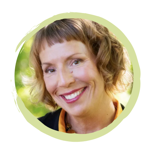 A woman smiling outdoors with short, curly blonde hair and wearing a dark jacket and orange shirt.