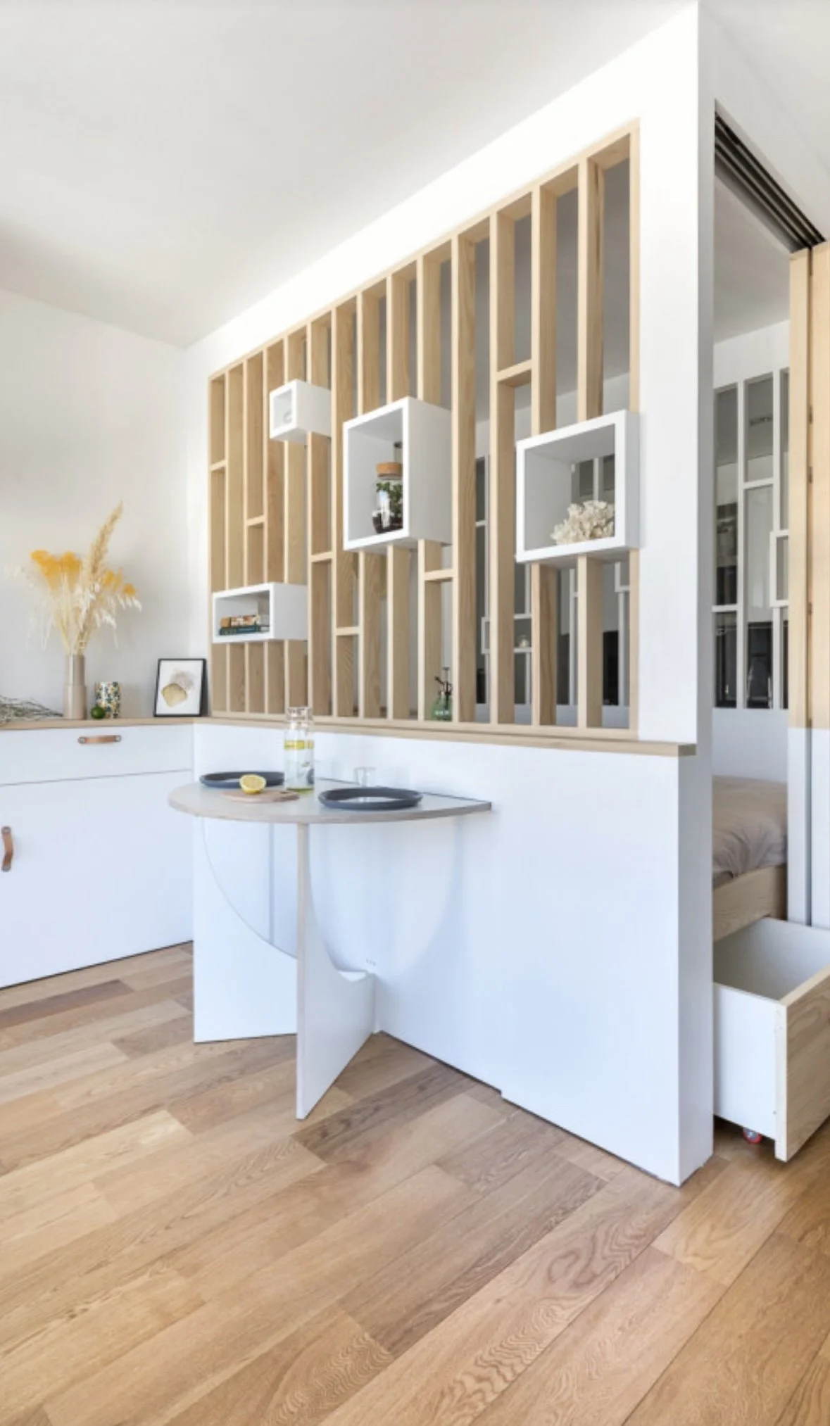 Intérieur d'un appartement avec une séparation en bois et des étagères blanches, une table ronde blanche avec deux assiettes et un citron, un tiroir en bois, une décoration florale en vase et un lit visible derrière une cloison.
