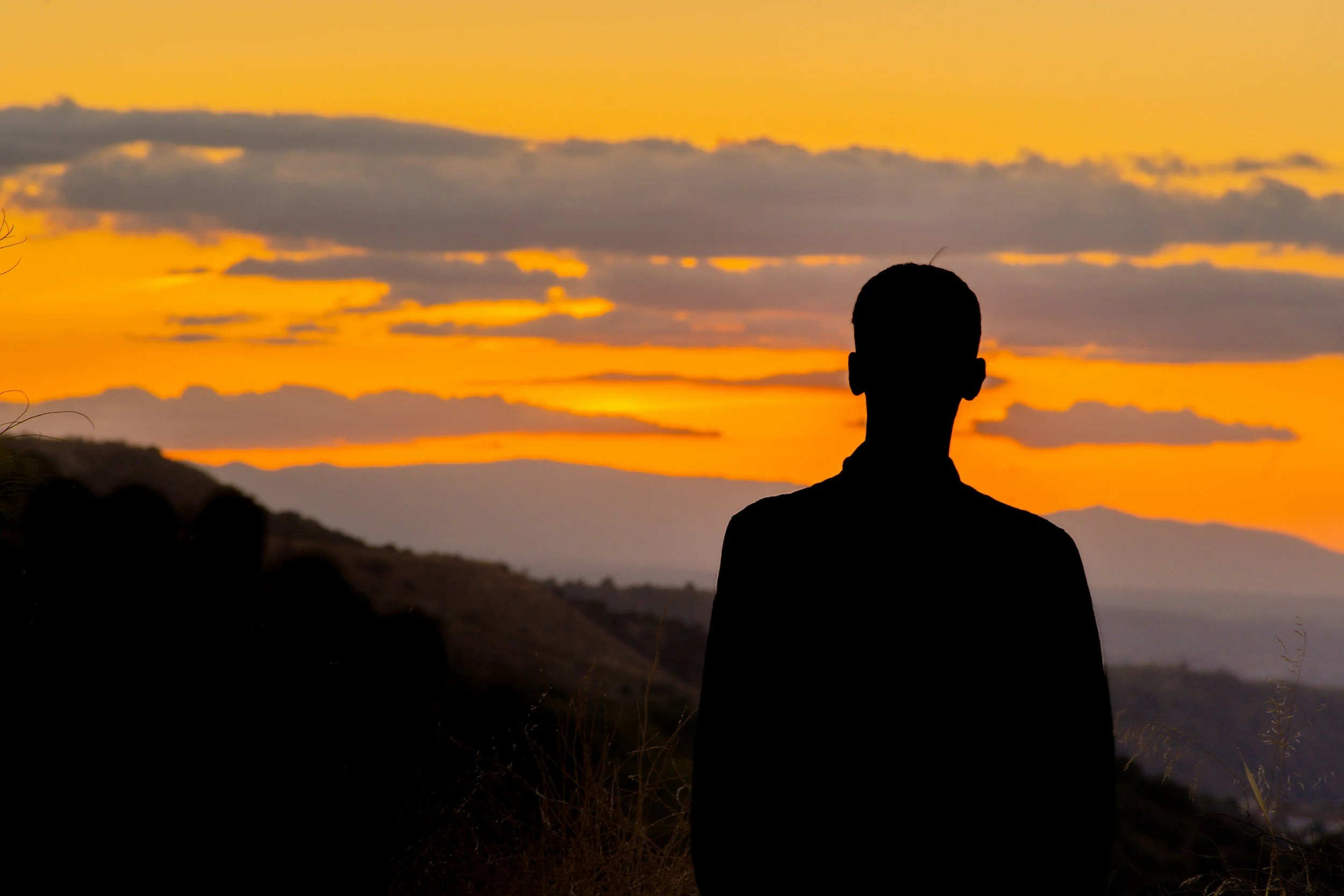 Silhouette of a person standing outdoors during sunset with a sky filled with orange and yellow hues and scattered clouds.
