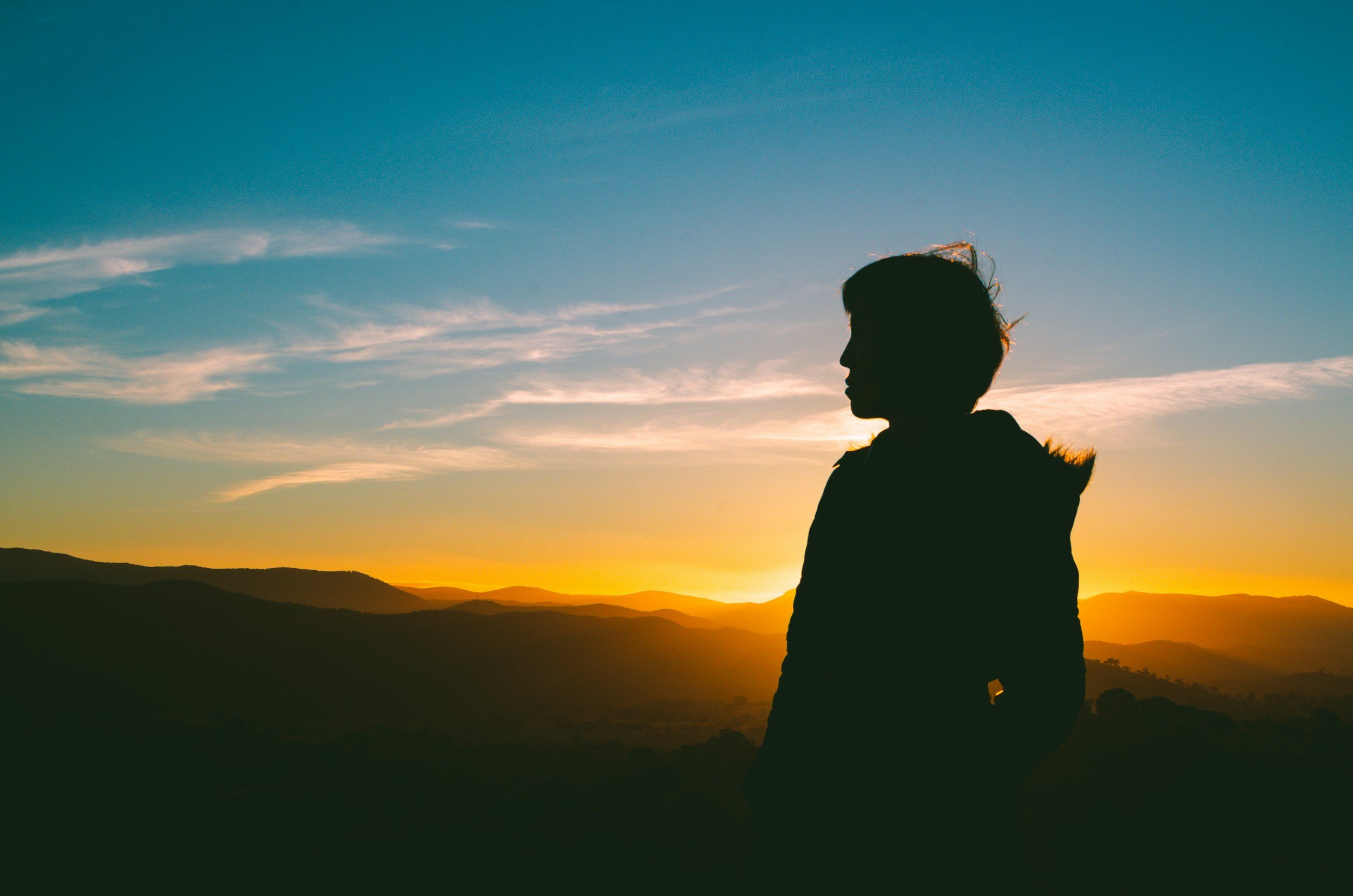 Silhouette of a person with short hair standing against a sunset over mountains and a blue sky with scattered clouds.