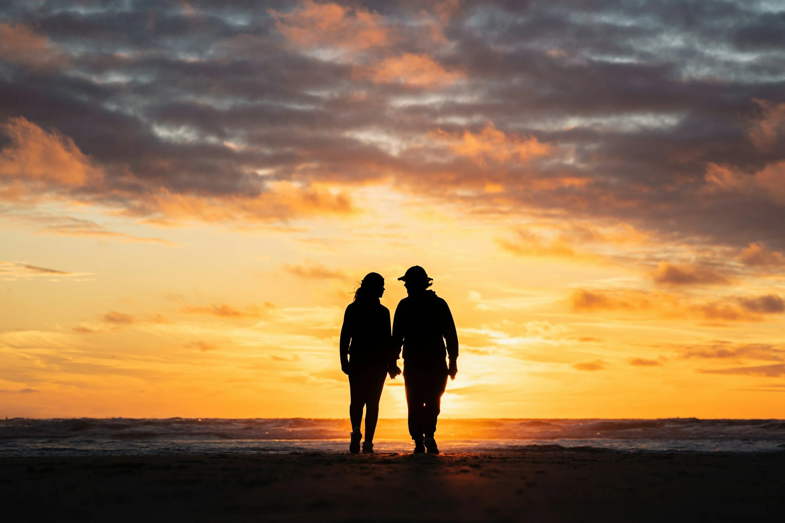 Silhouettes of a couple holding hands and walking on the beach during sunset, with a colorful sky filled with clouds.