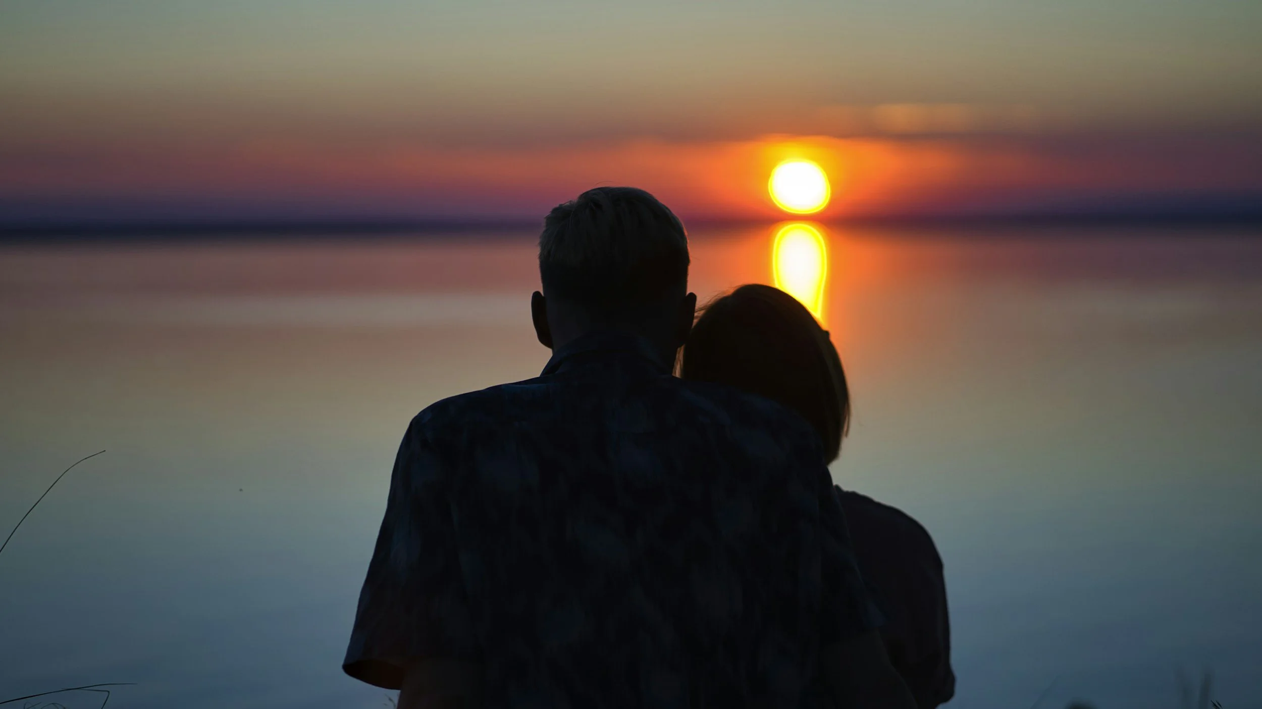 Couple sitting by a calm body of water during sunset, with the sun low on the horizon and colorful sky.