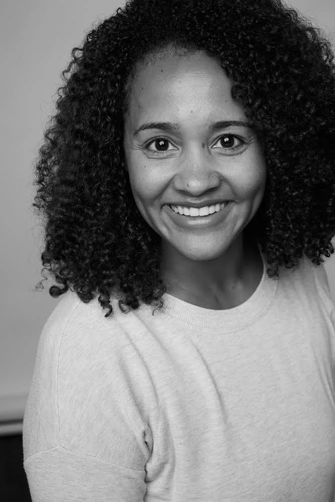 Black and white photo of a woman with curly hair smiling at the camera. She is wearing a light-colored T-shirt.