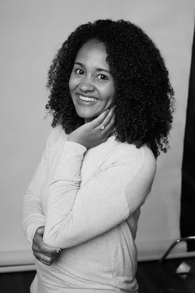 Black and white portrait of a woman with curly hair, smiling, holding her chin with one hand, wearing a long-sleeve shirt.