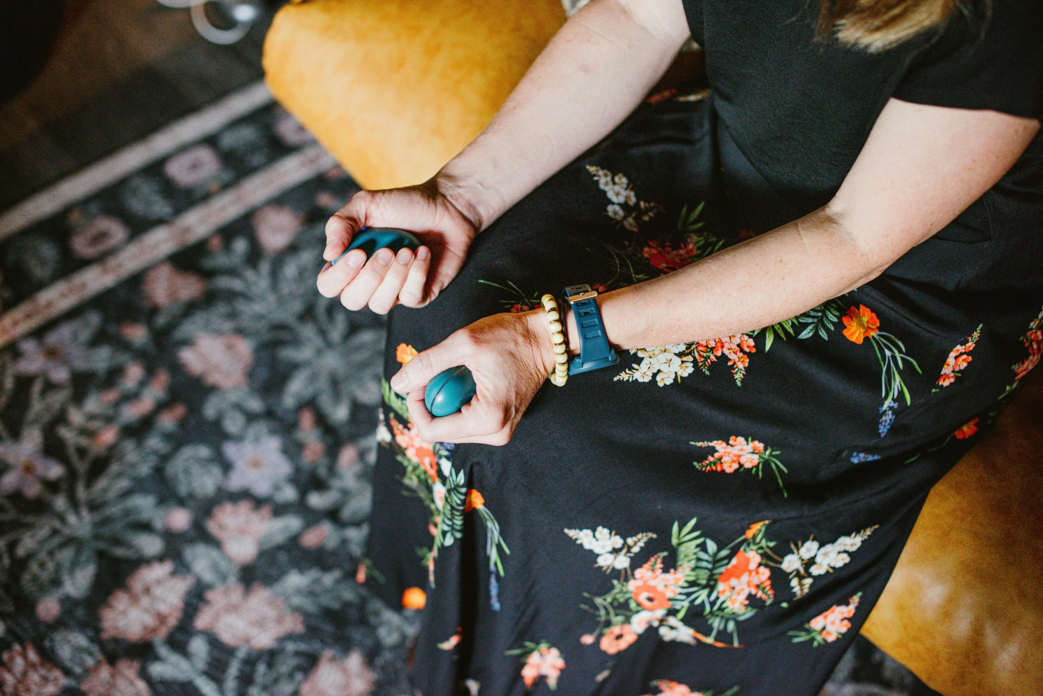 A woman sitting on a yellow cushion holding two computer mice, wearing a black dress with floral pattern, a blue smartwatch, and a yellow beaded bracelet.