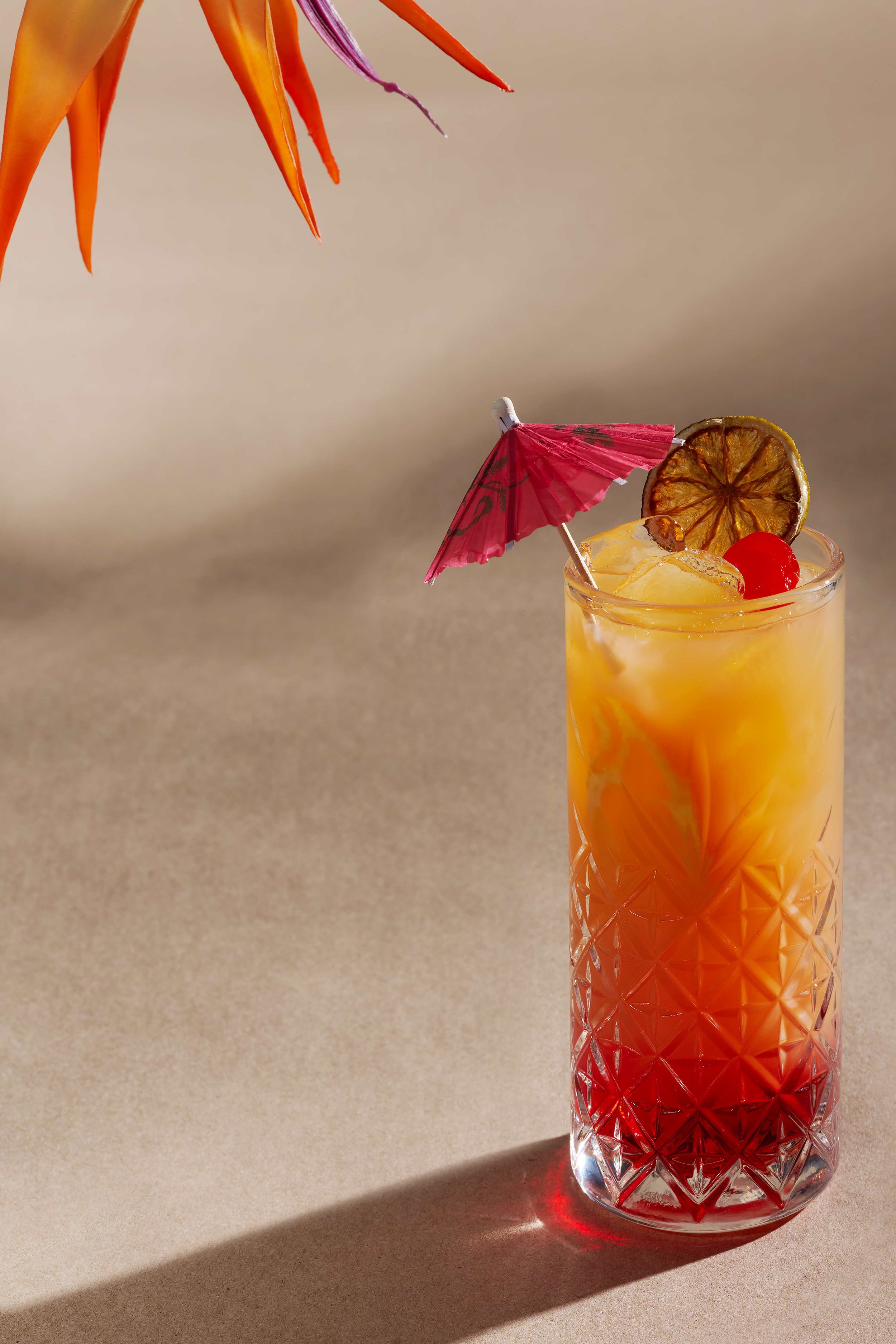 Tropical tequila sunrise cocktail in a crystal highball glass with a red umbrella, maraschino cherry, and dried lime garnish on a sandy background
