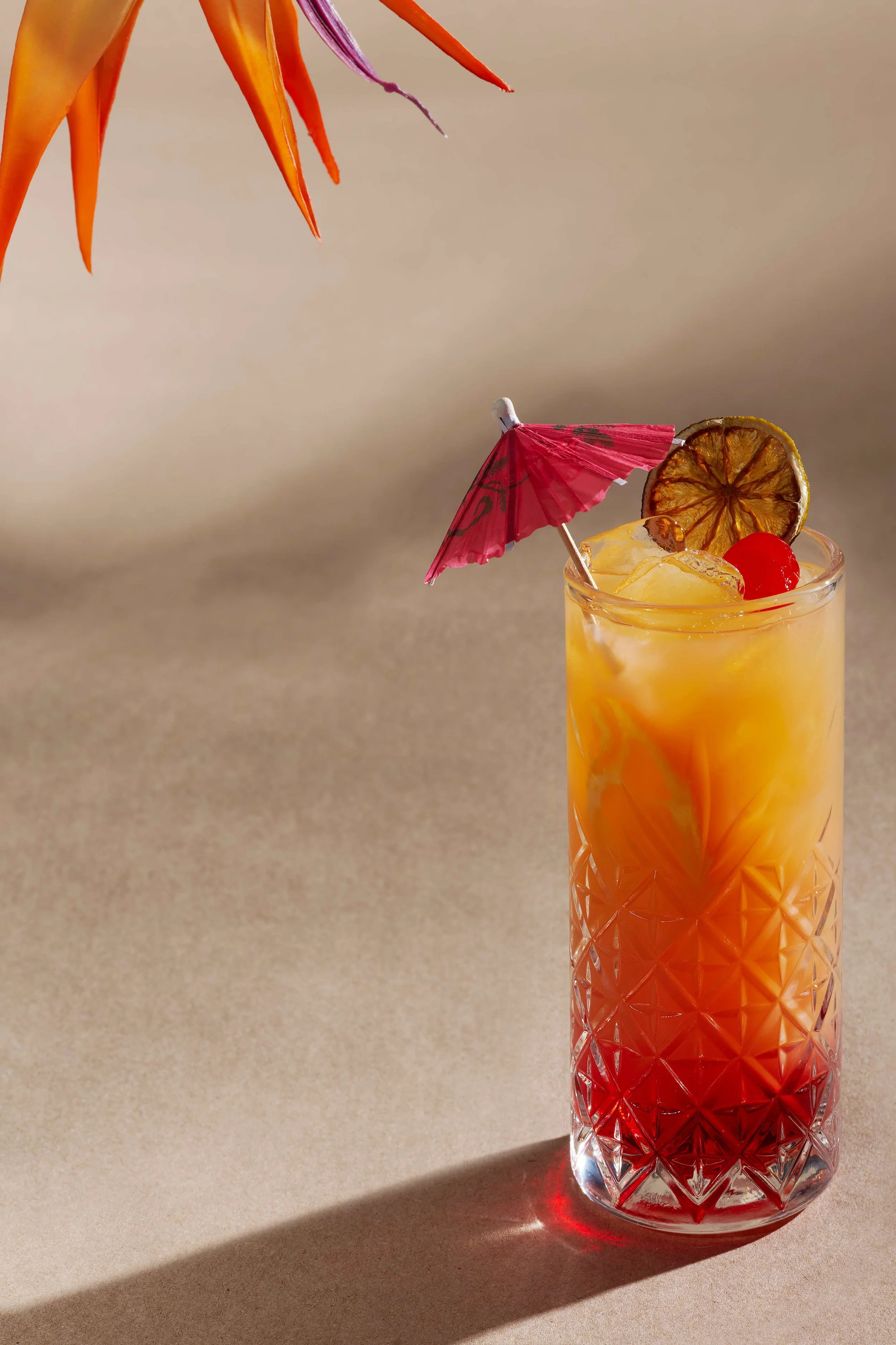 Tropical tequila sunrise cocktail in a crystal highball glass with a red umbrella, maraschino cherry, and dried lime garnish on a sandy background