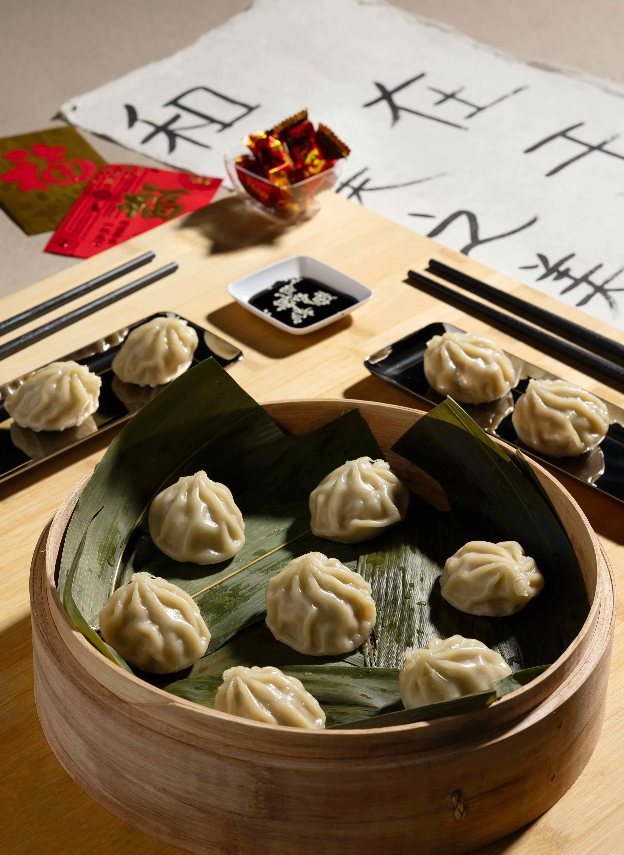 Steaming soup dumplings (Xiao Long Bao) in a bamboo steamer basket lined with banana leaves, arranged on a wooden table with calligraphy, red envelopes, and soy dipping sauce