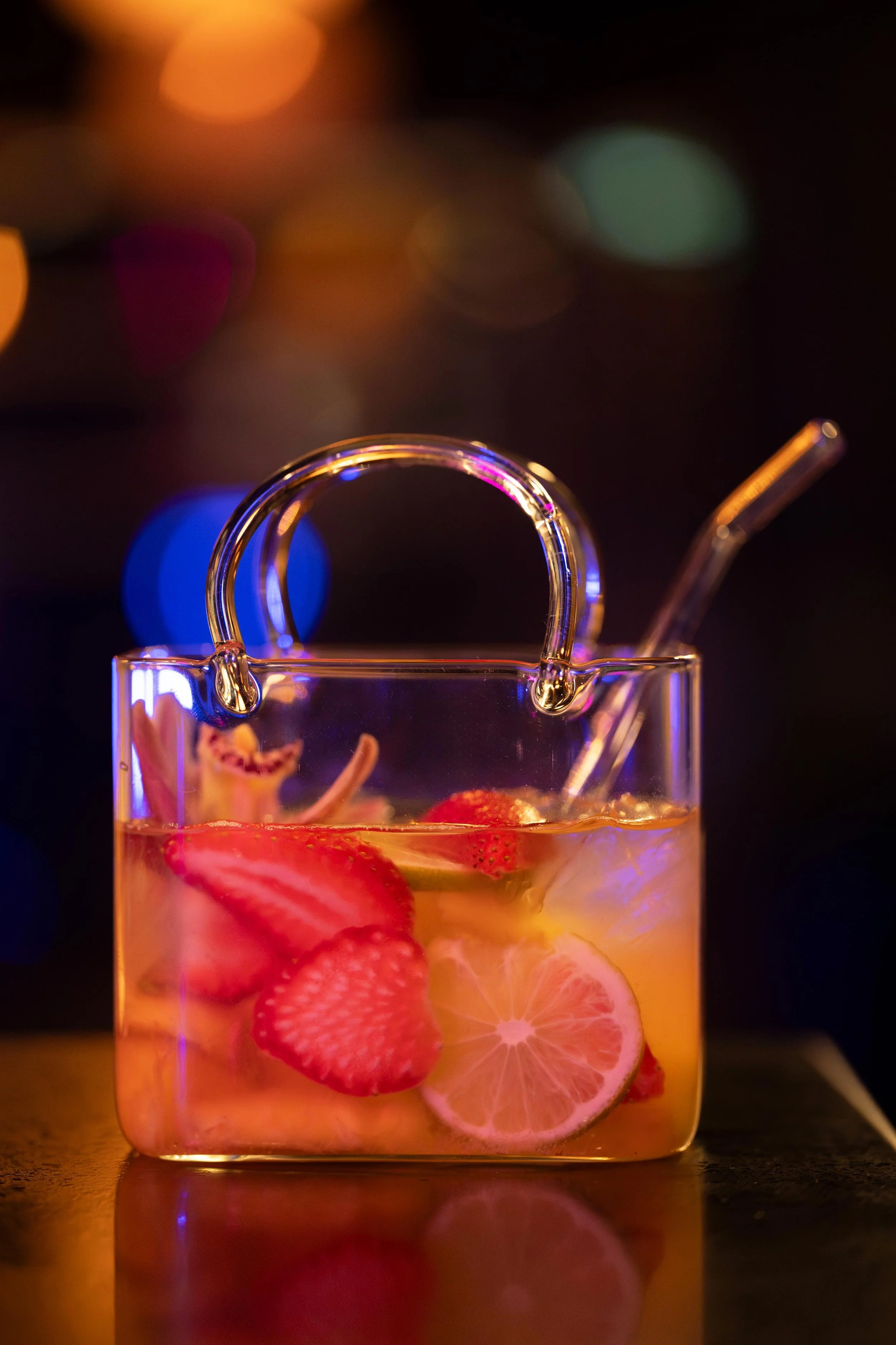 Unique Bolsita cocktail served in a glass purse-shaped mug with fresh strawberry slices, lime, and a glass straw against a bokeh night background