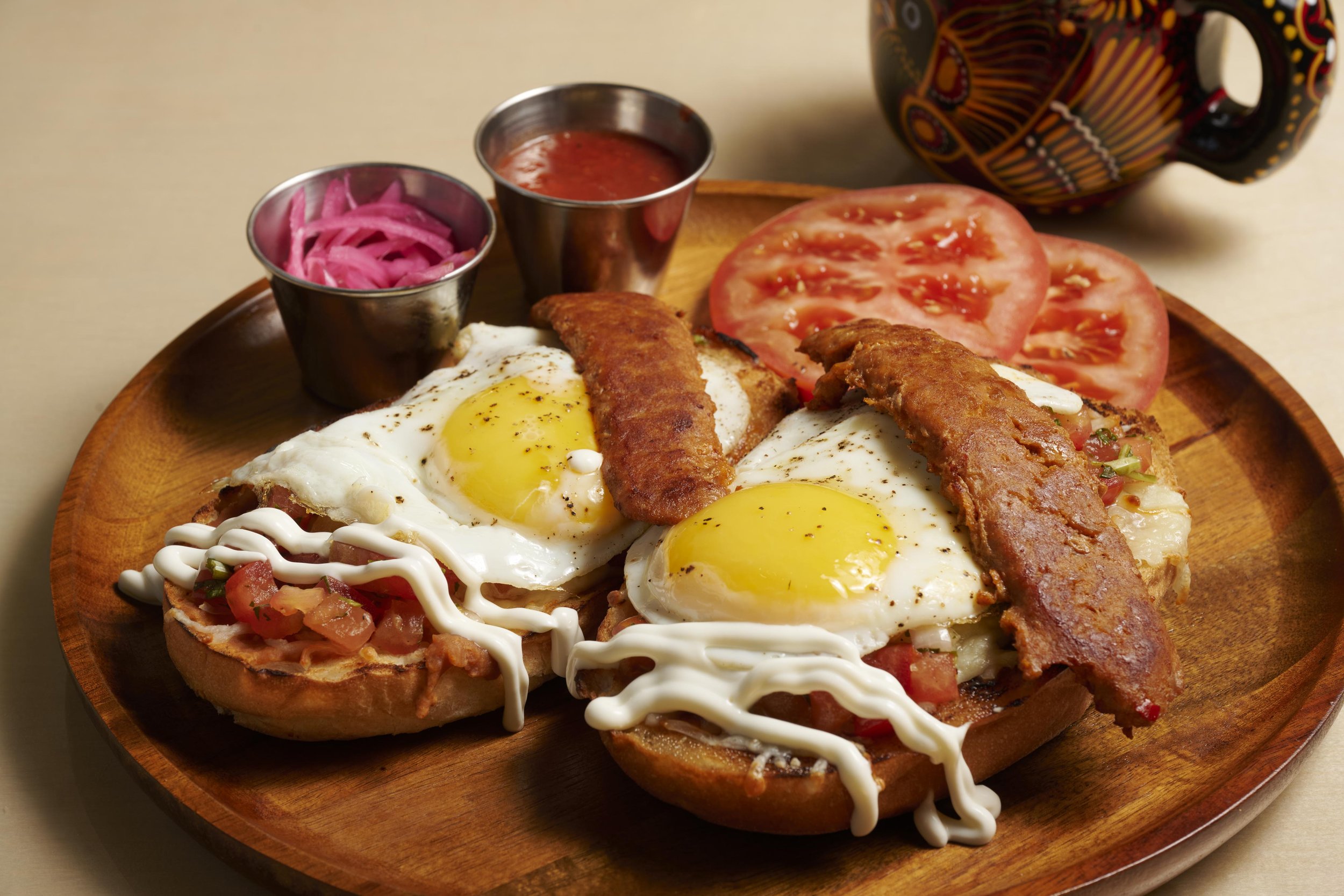 Mexican breakfast molletes topped with fried eggs, crispy bacon, pico de gallo, and crema, served on a wooden plate with pickled onions and salsa