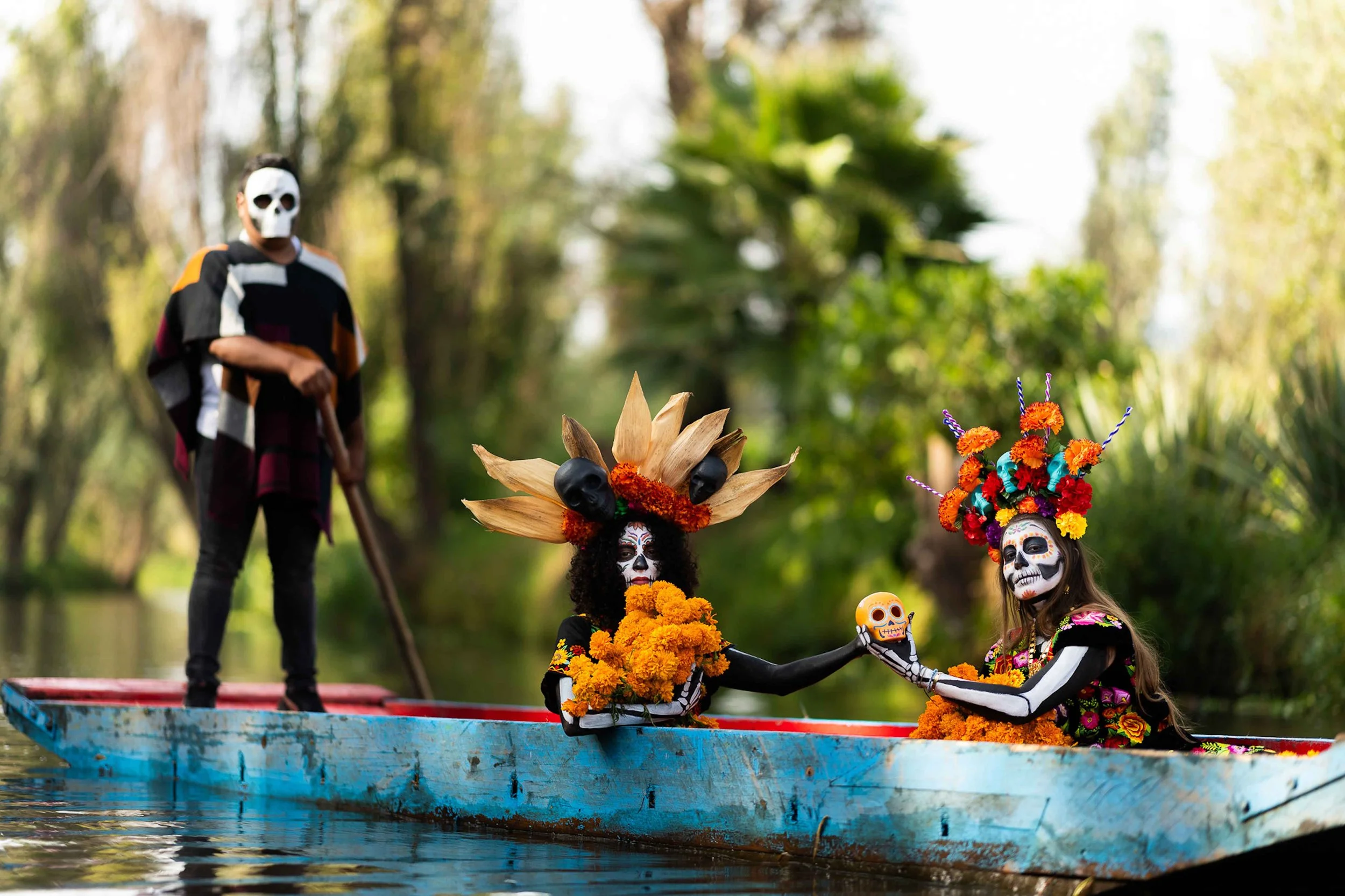 Two women in Catrina skull makeup and traditional Day of the Dead costumes sitting in a blue trajinera boat on a canal in Xochimilco, Mexico.