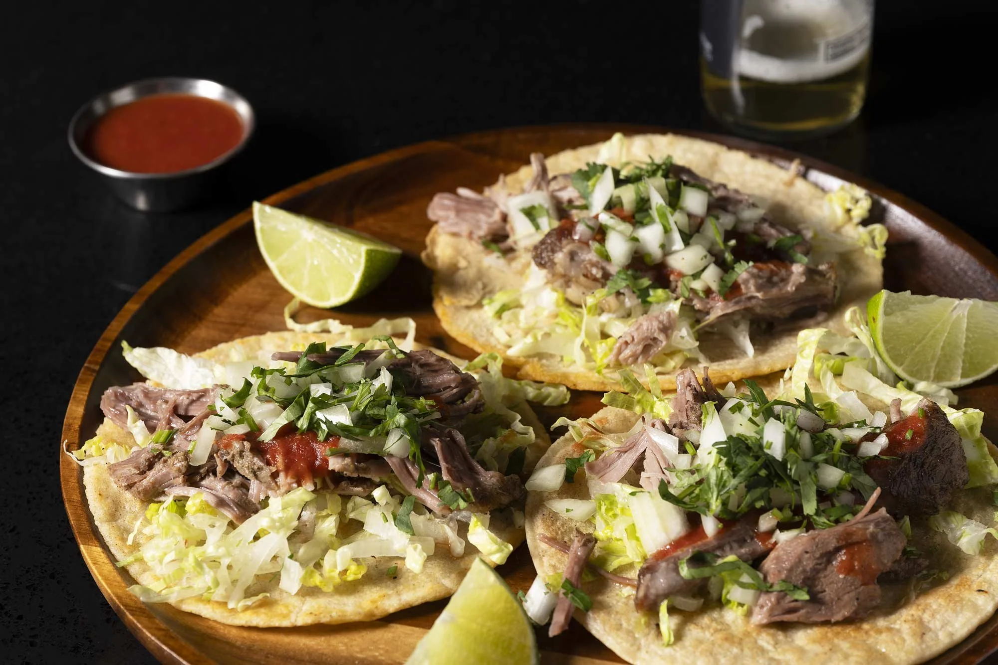 Three authentic pork carnitas tacos on corn tortillas with shredded cabbage, onion, cilantro, and red salsa, served on a wooden plate with lime wedges