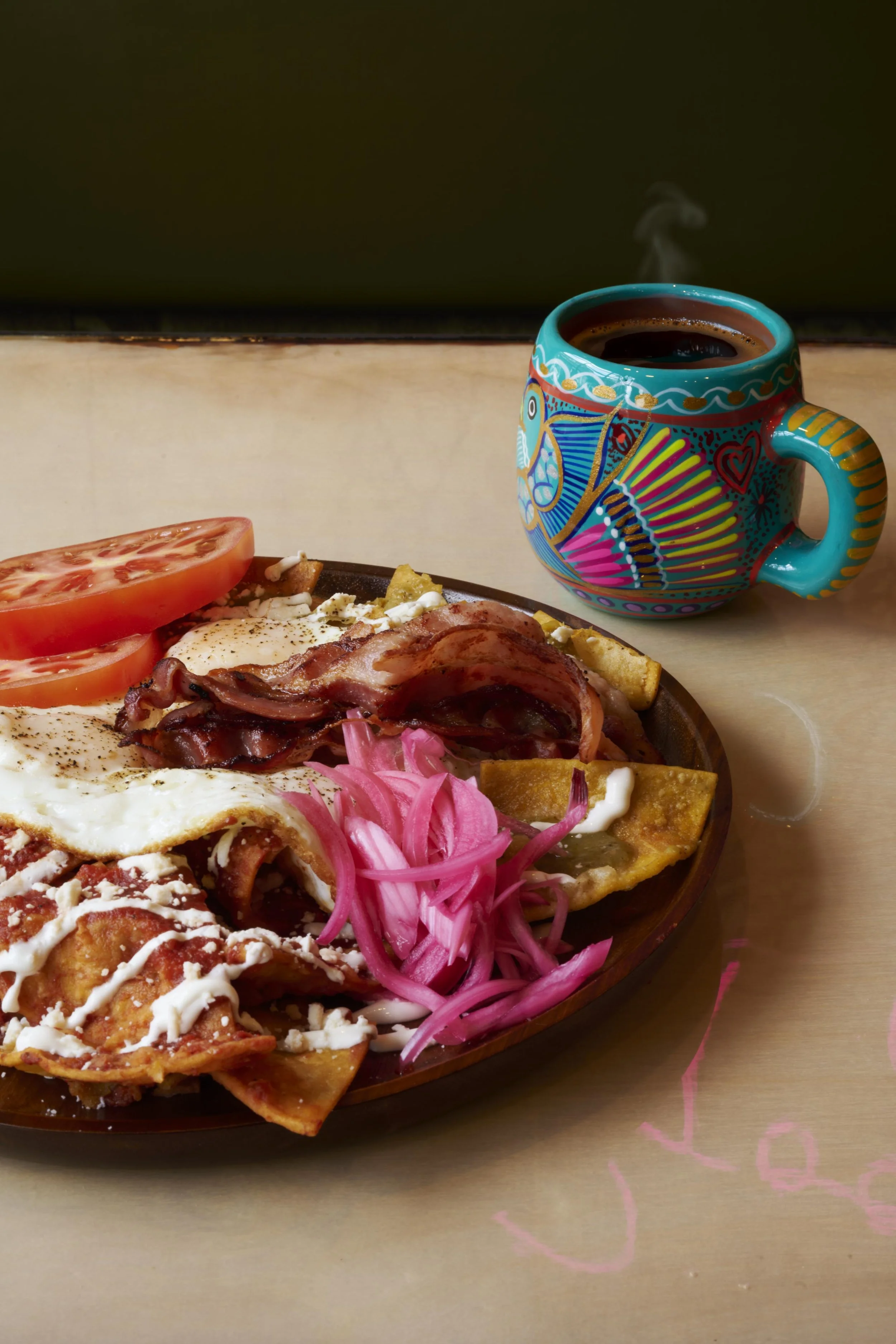 Plate of Mexican chilaquiles with fried eggs, crispy bacon, pickled onions, and tomatoes served next to a steaming cup of coffee in a hand-painted turquoise talavera mug