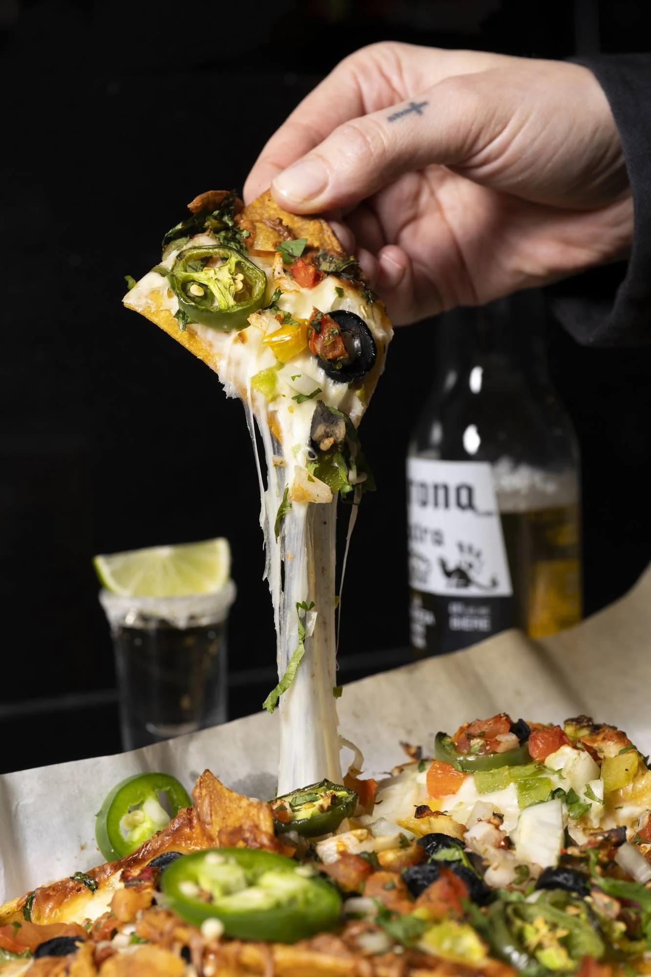 Close-up of a hand lifting a loaded nacho with long melted cheese strings, jalapeños, olives, and pico de gallo, served with a Corona beer and tequila shot