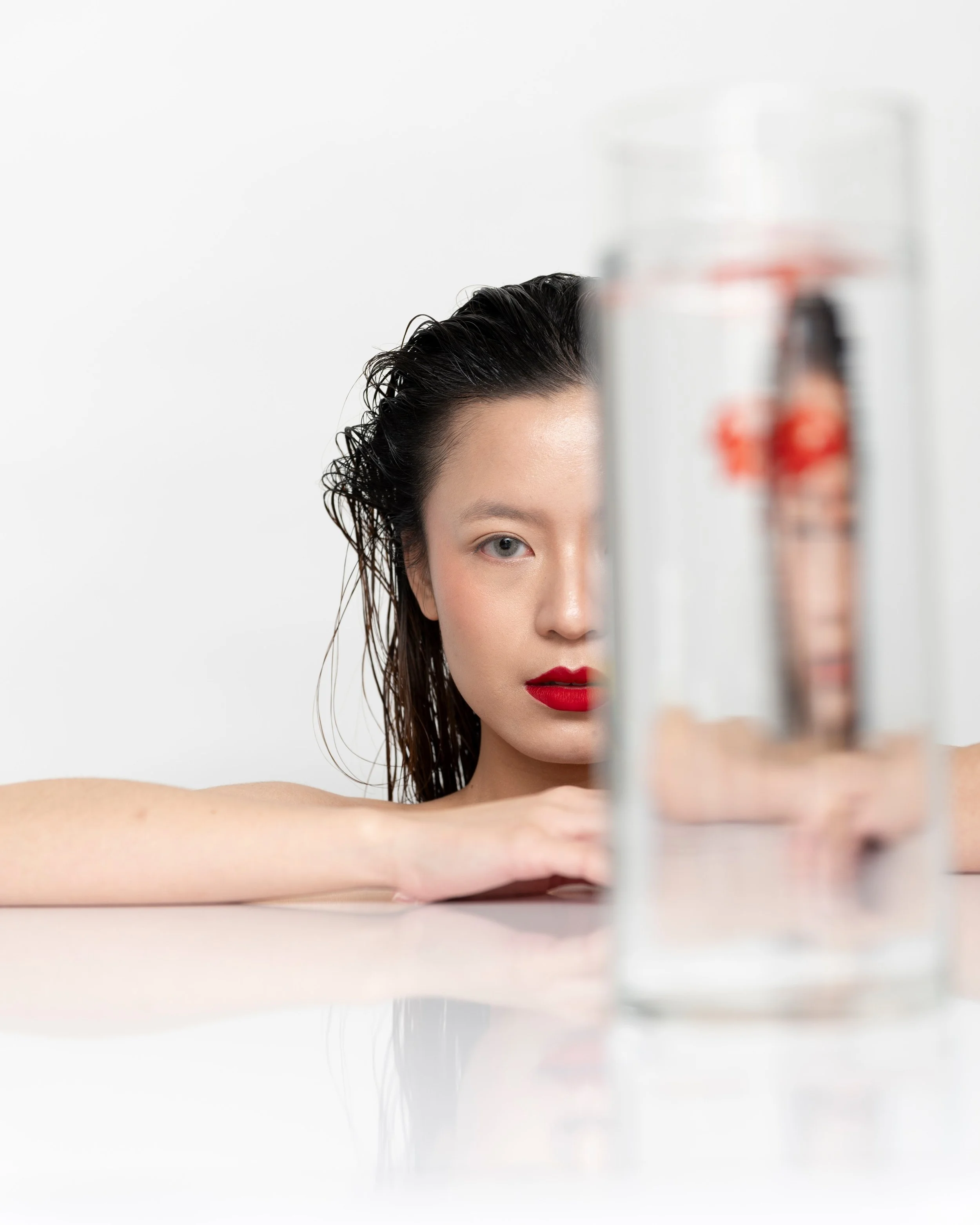 Close-up of a woman with wet hair and bold red lipstick looking through a tall glass of water, creating a distorted reflection on a clean white background