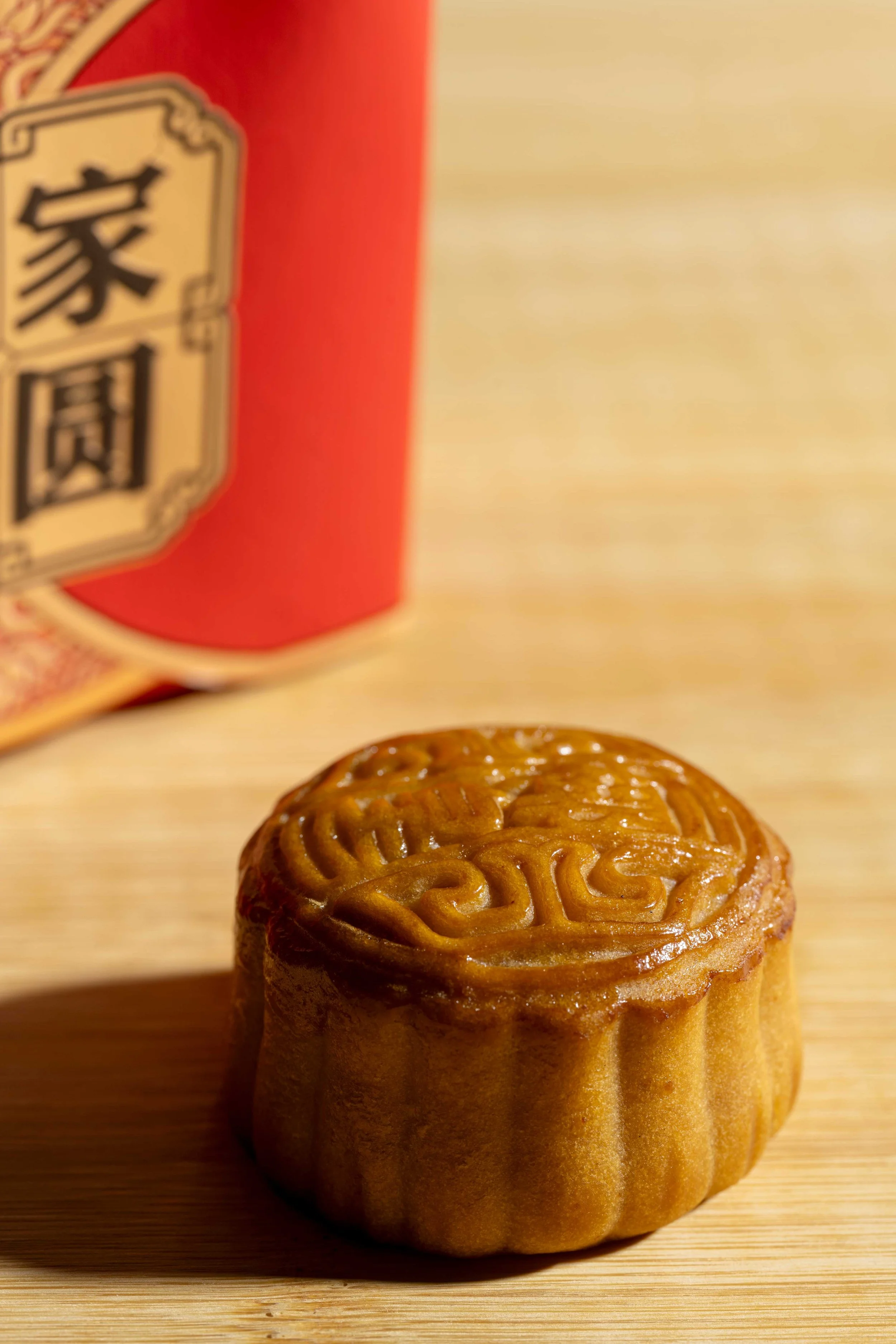 A traditional Chinese mooncake with an intricate pattern on a wooden surface, featuring a red gift box with Chinese calligraphy in the blurred background