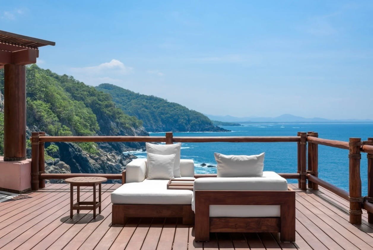 A wooden deck with white cushioned seating and a small round table overlooks a scenic ocean view with green hills and a blue sky.