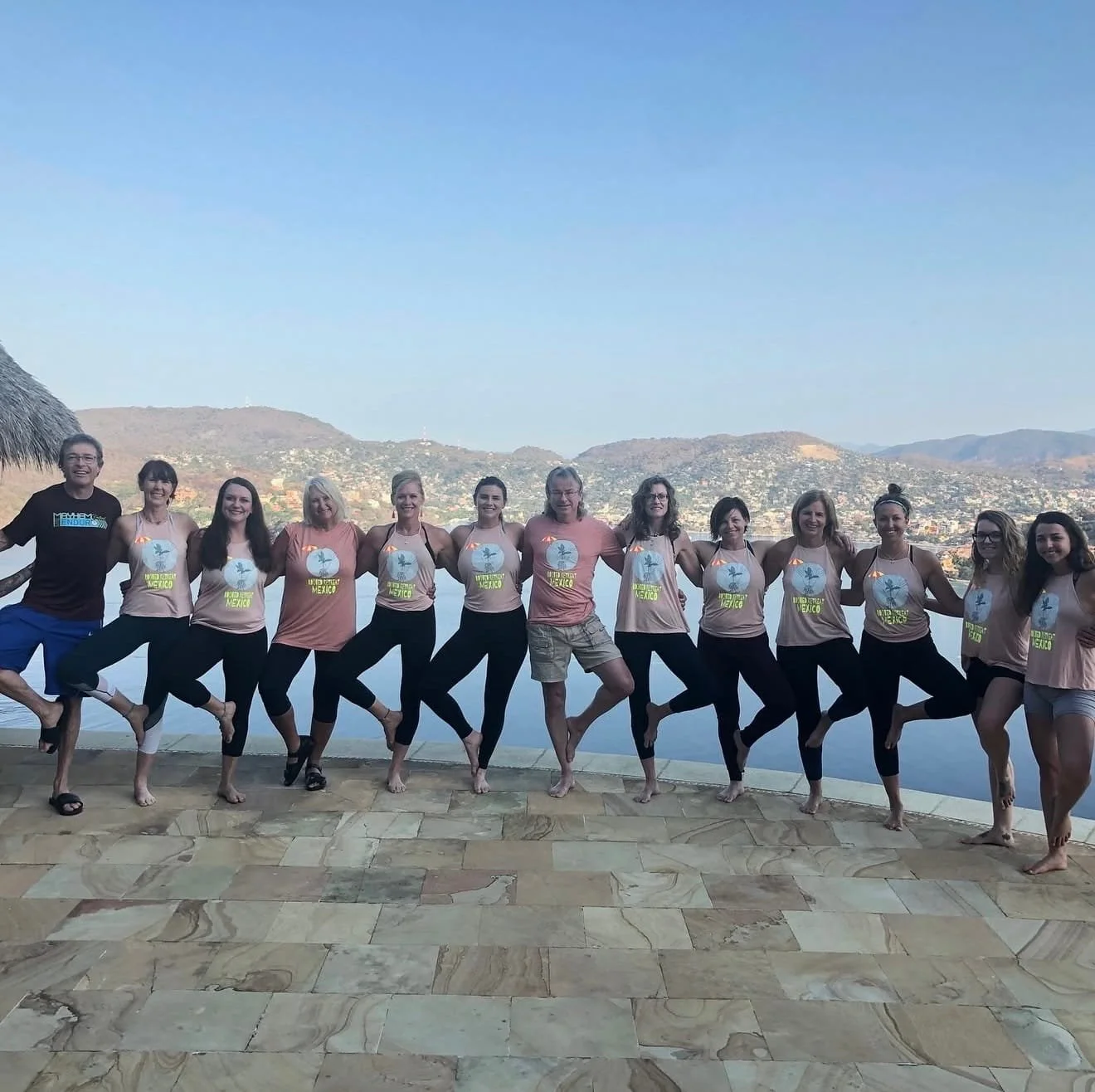 Group of people standing side by side on a balcony, outdoors, with mountains and city in the background, smiling, dressed in casual athletic clothes.