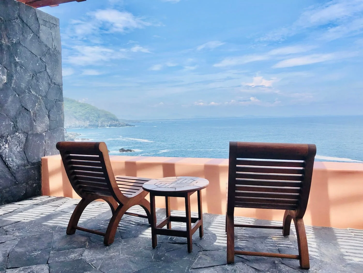Two wooden lounge chairs and a small table on a stone patio overlooking the ocean with blue sky and clouds.