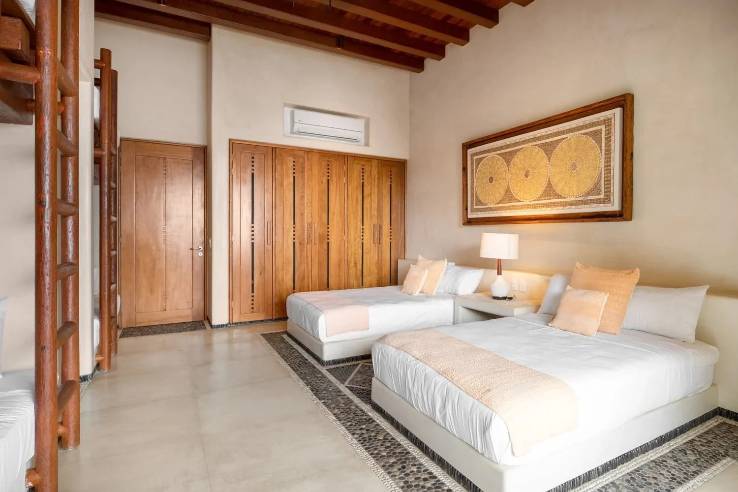 A bedroom with two white beds, beige pillows, a wooden headboard, a single bedside table with a lamp, wooden closet doors, beige walls, a framed wall art piece, and beige tiled flooring.