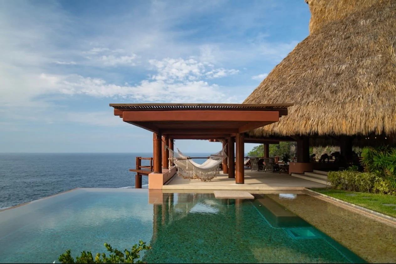 A luxury seaside villa with an infinity pool, a hammock under a wooden structure, and a thatched roof over an open-air lounge area, with ocean and blue sky in the background.