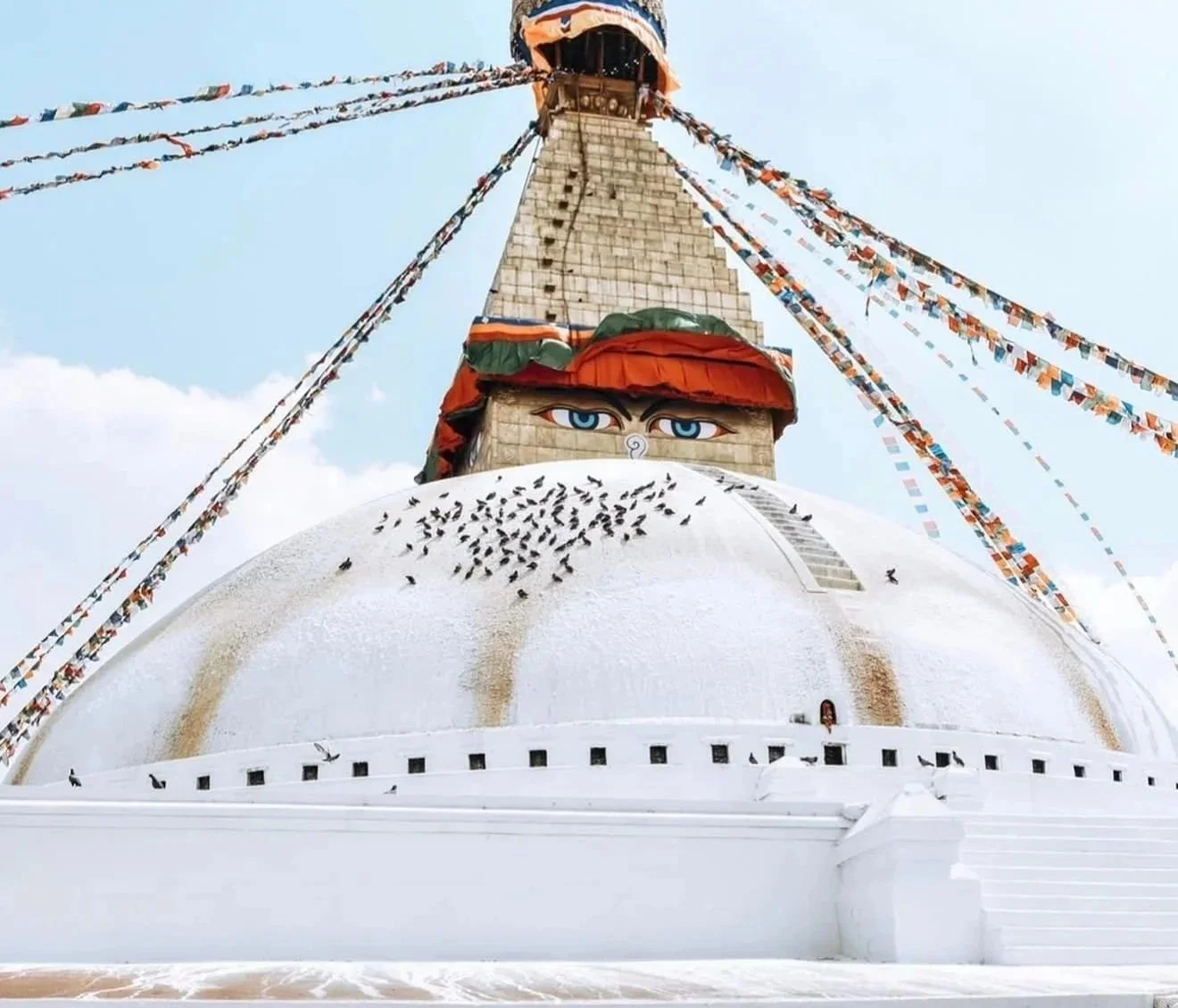 A white stupa with a large pair of painted blue eyes above the entrance, decorated with colorful prayer flags extending from the top, and birds perched on its surface.