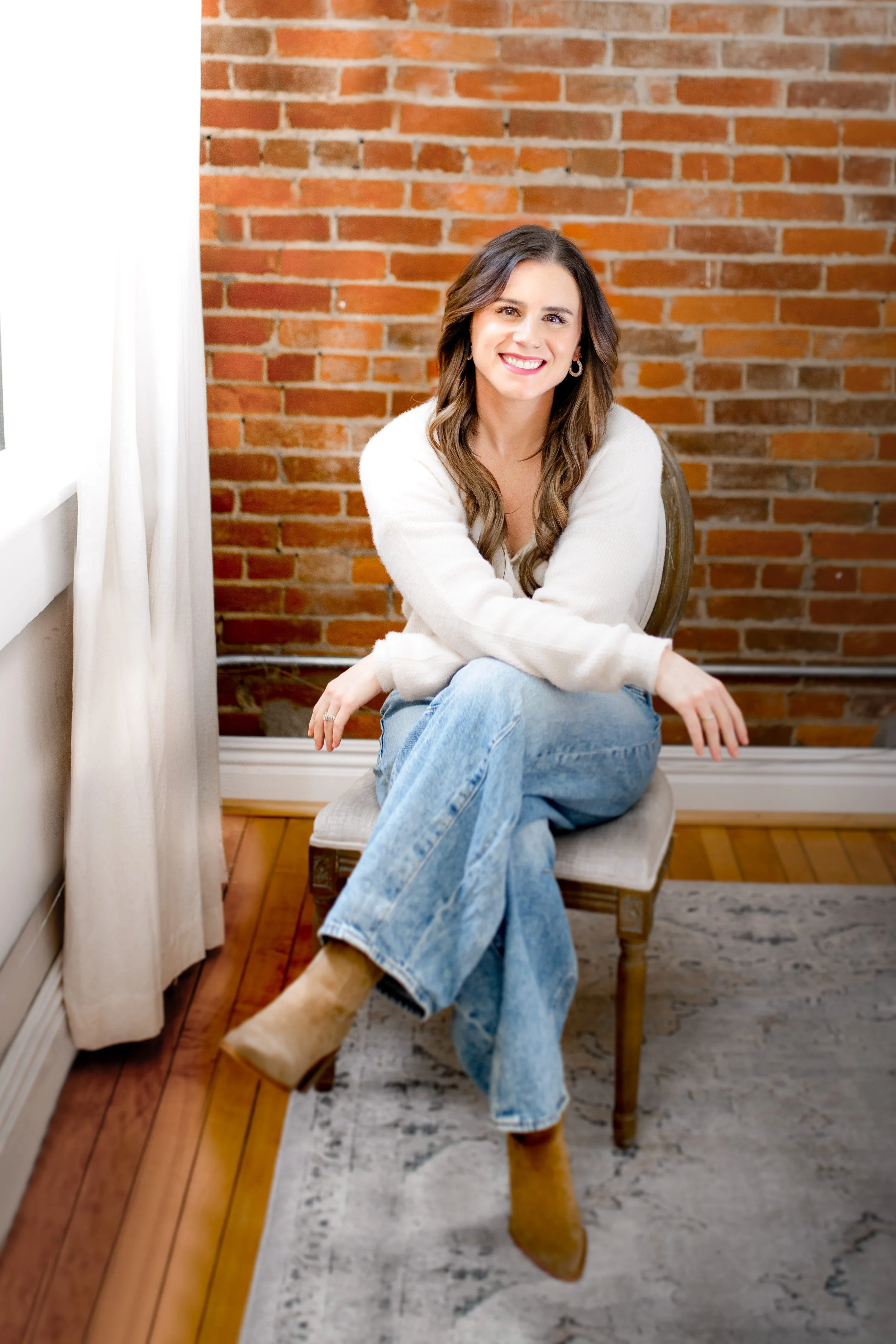 A woman with long wavy brown hair, wearing a white sweater, blue jeans, and tan boots, sitting on a beige chair in a room with a brick wall and a window with white curtains.