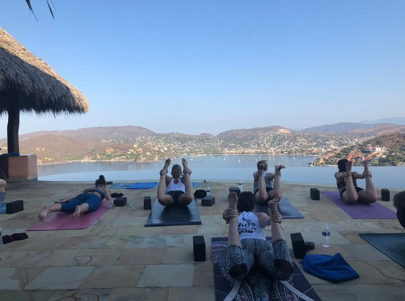People practicing yoga outdoors on mats near a body of water with hills and a town in the background.
