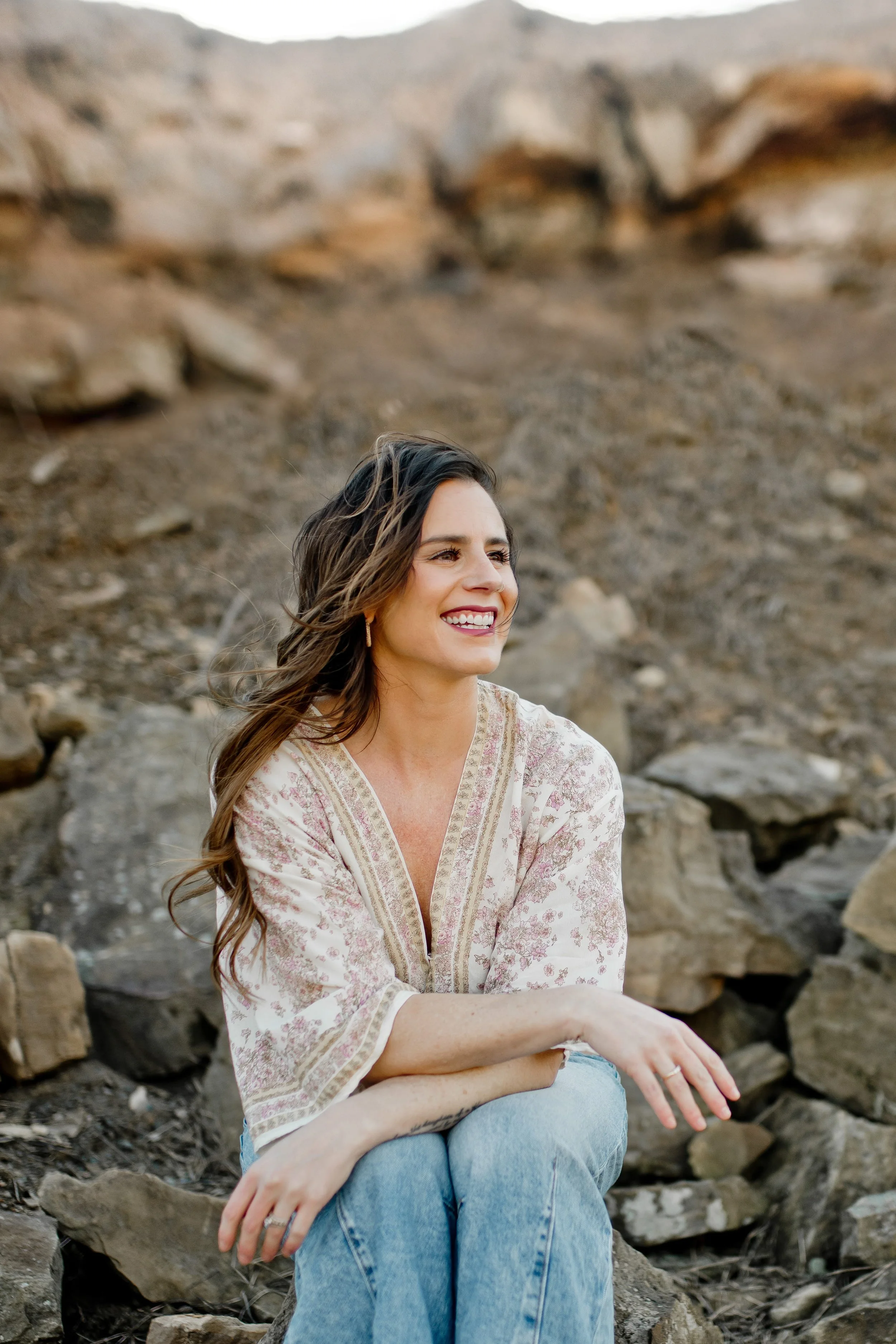 A woman sitting outdoors on rocks, smiling, with a rocky hillside background.