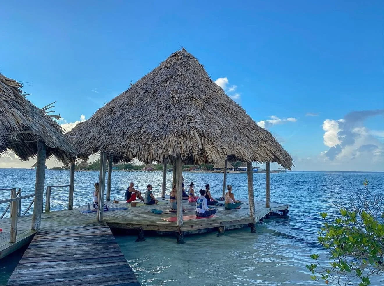 People practicing yoga on a wooden platform under thatched huts over water, with a clear blue sky.
