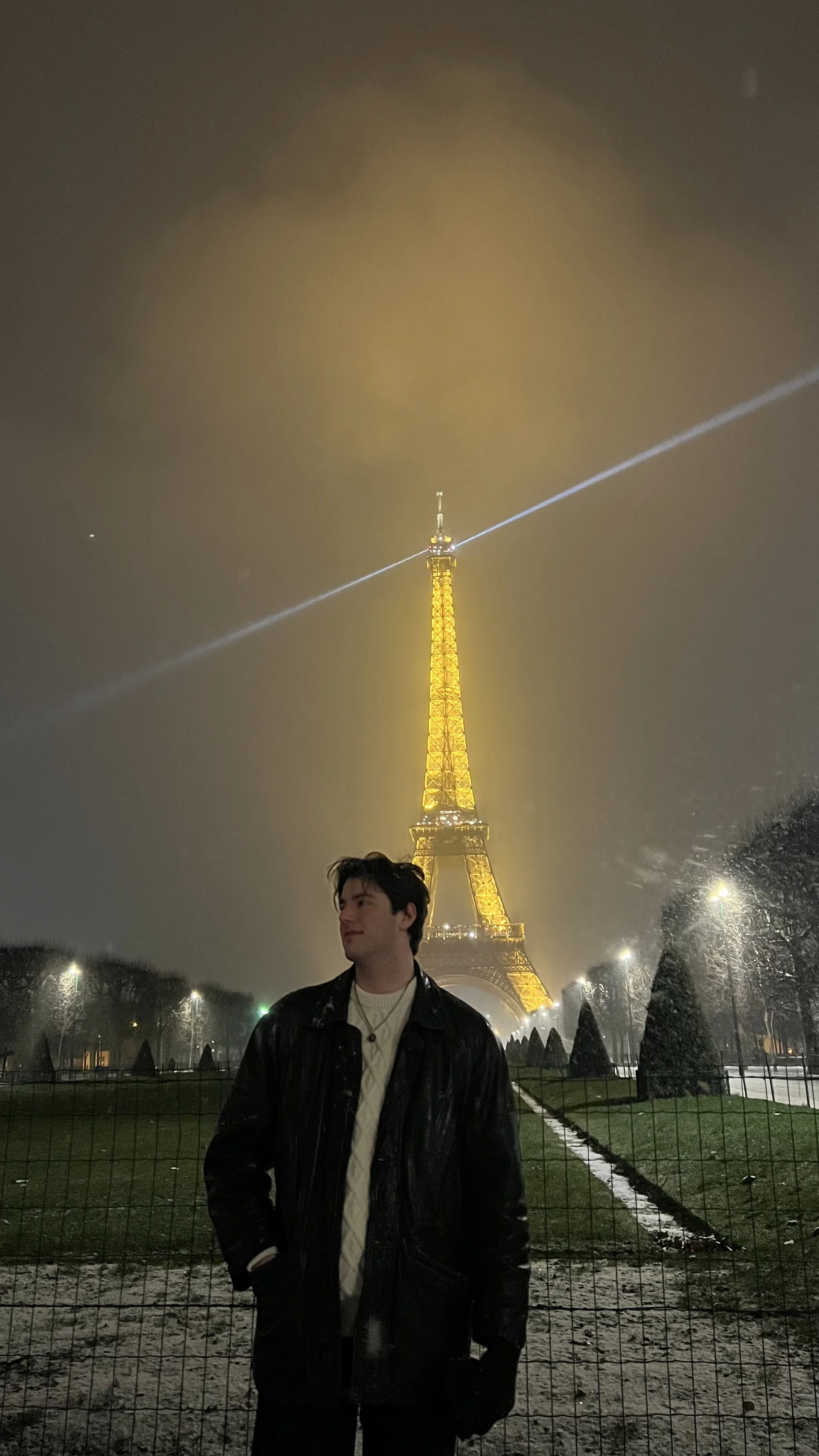 A young man standing outdoors at night in front of the illuminated Eiffel Tower, with foggy weather and snow on the ground.