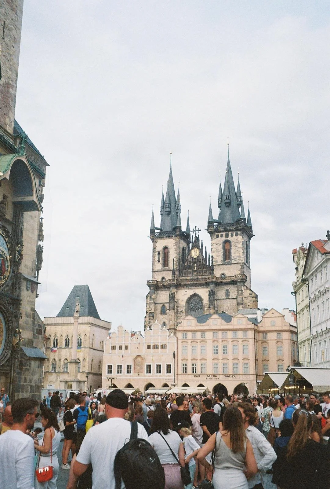 Crowd of people gathered in a town square with historic buildings and a Gothic-style church with twin spires in the background.