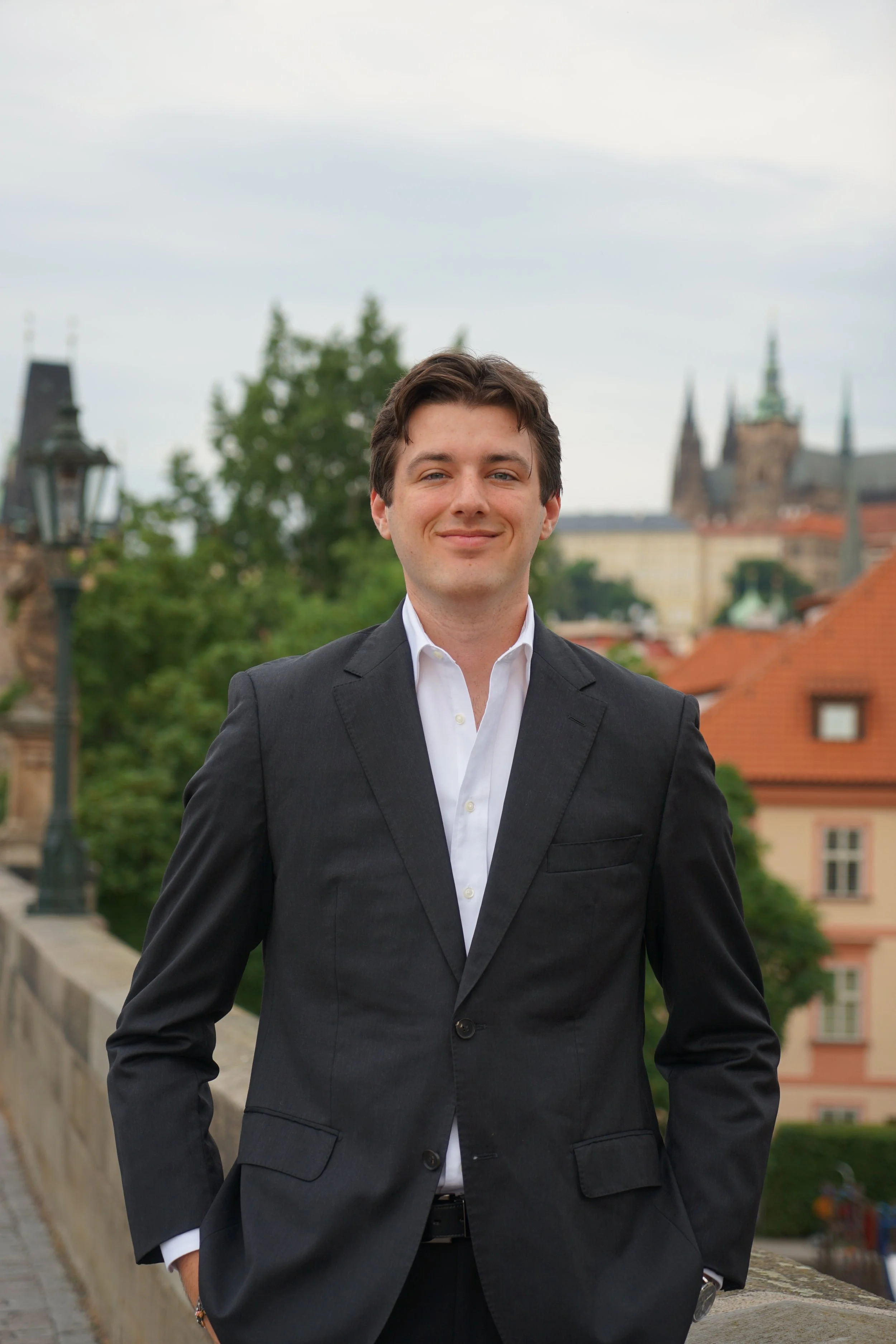 A young man wearing a black suit jacket and white shirt standing outdoors on a bridge with a historic cityscape and church spires in the background.