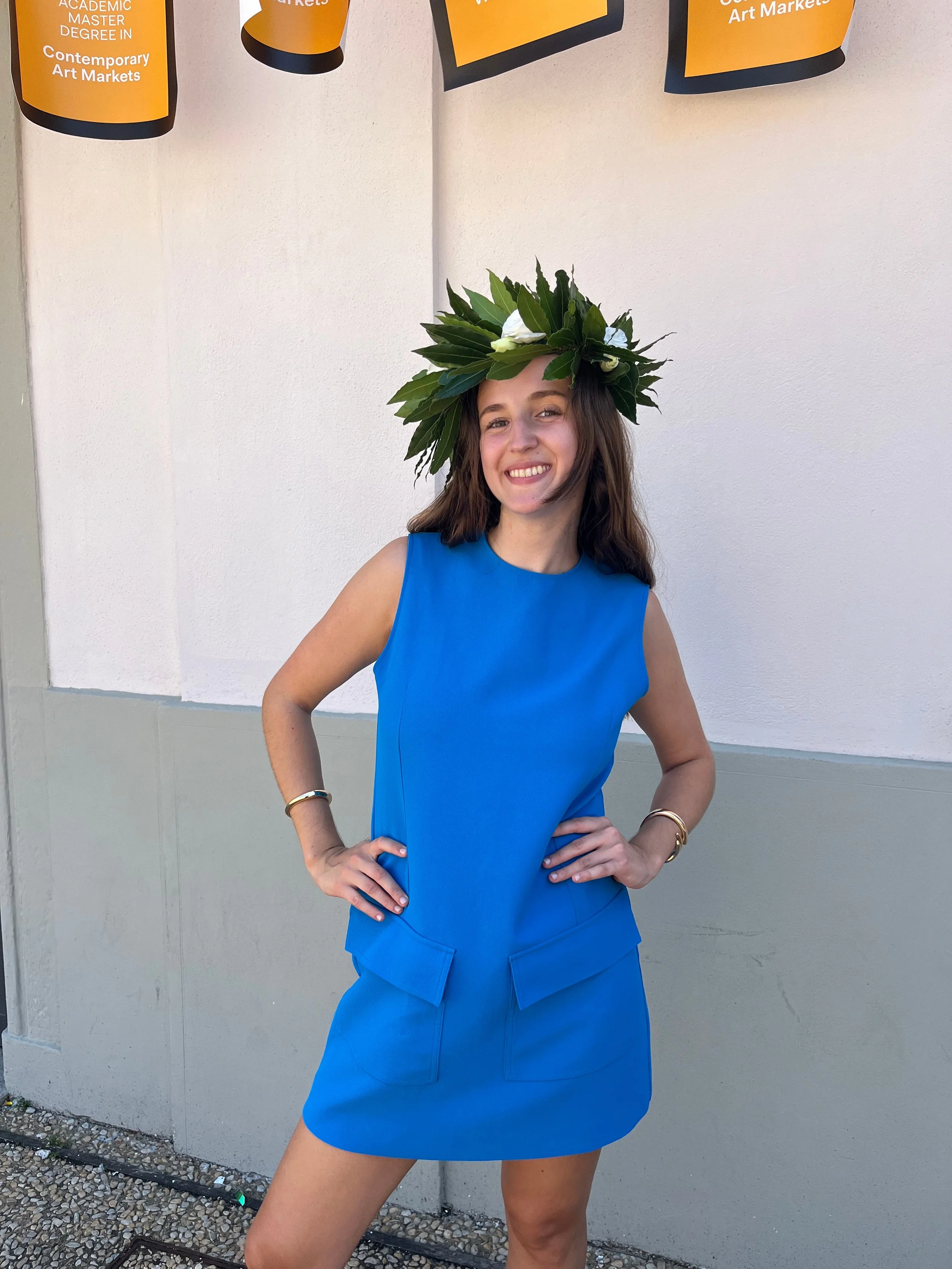 A young woman wearing a blue sleeveless dress and a leafy crown, standing outdoors with hands on hips, smiling, and posing in front of a white wall.