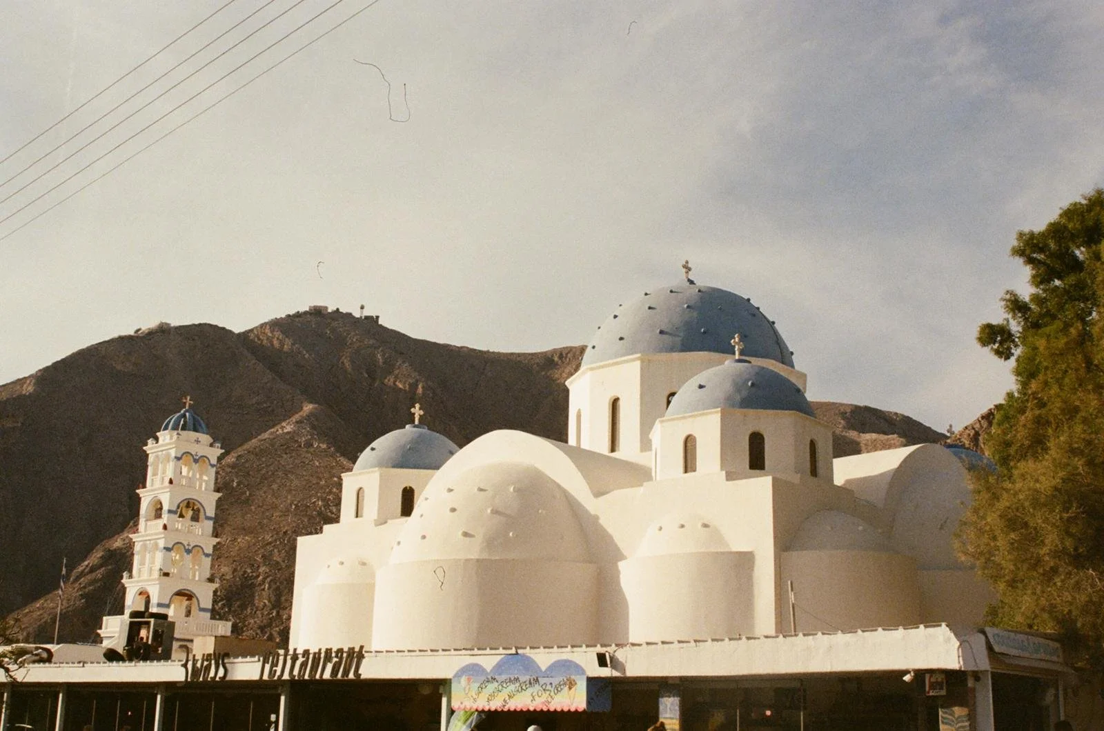 White Greek Orthodox church with blue domes and crosses, mountain in the background, trees on the right, and a sign in front.