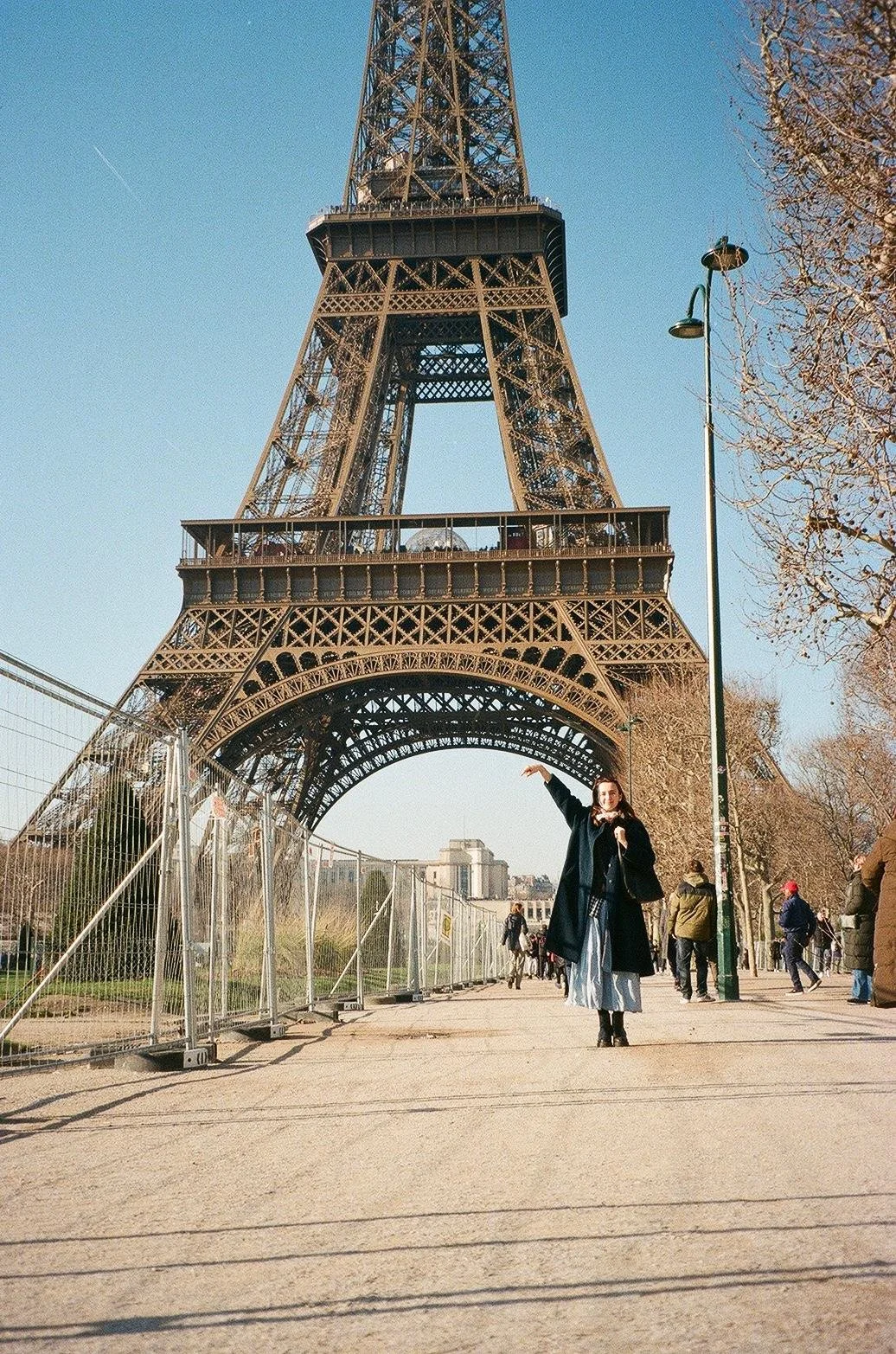 A woman standing in front of the Eiffel Tower in Paris, France, pointing upwards with her right hand, with other visitors and trees in the background on a clear day.