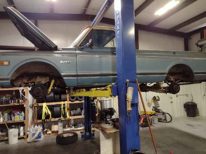 A vintage blue pickup truck raised on a hydraulic lift in a garage or workshop, with the front wheel removed.
