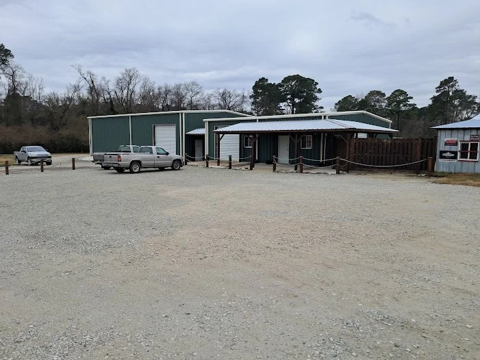 A gravel parking lot with three parked vehicles in front of a green metal building and a smaller blue structure, surrounded by wooden posts and rope barriers, with trees and overcast sky in the background.