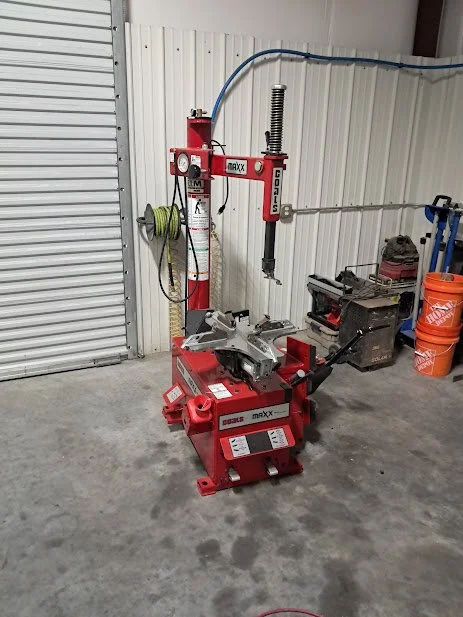 A red tire changer machine in a workshop garage, with a metal wall, a roll-up door, and orange buckets in the background.