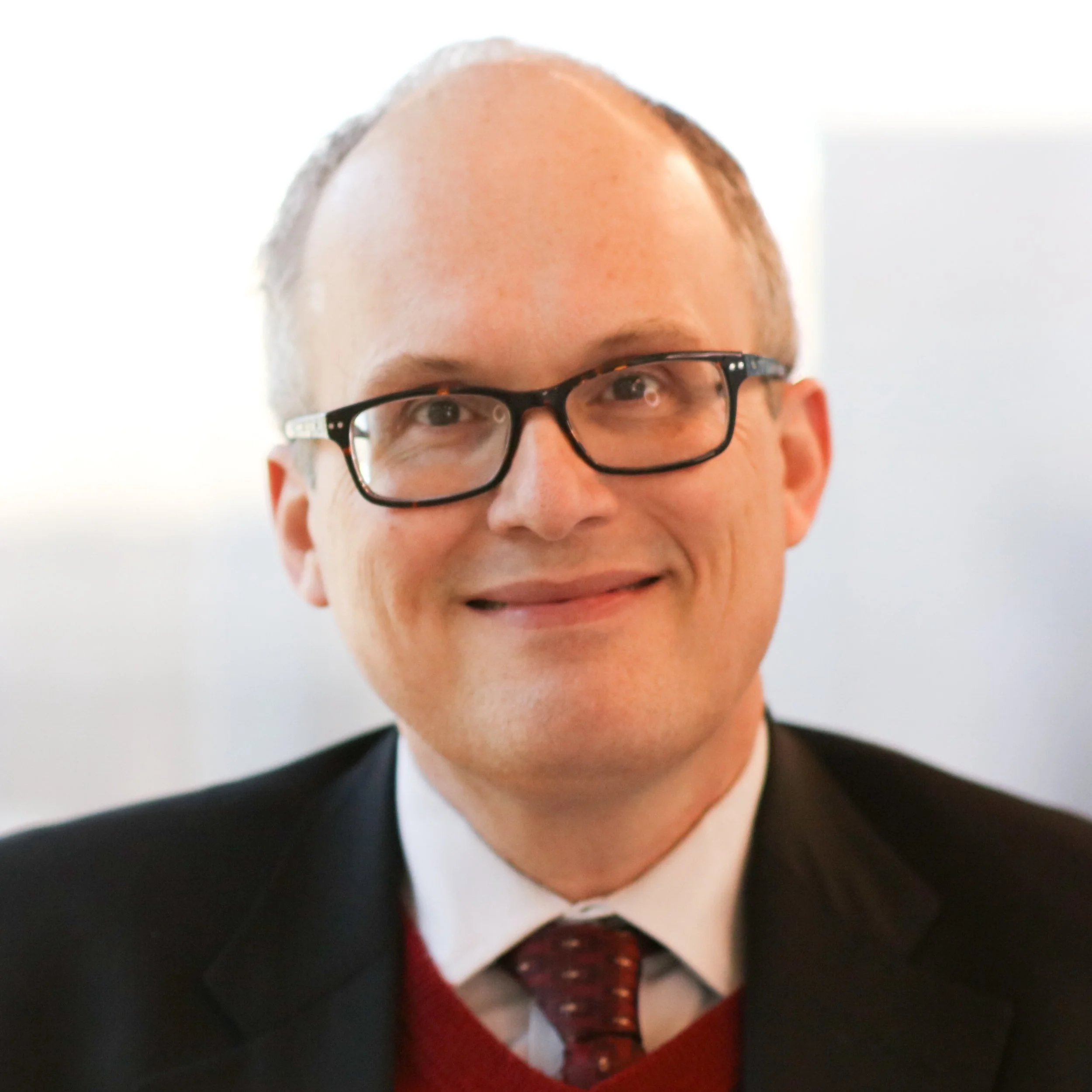 Close-up of a man with glasses and a red tie, wearing a black suit and white shirt, smiling.