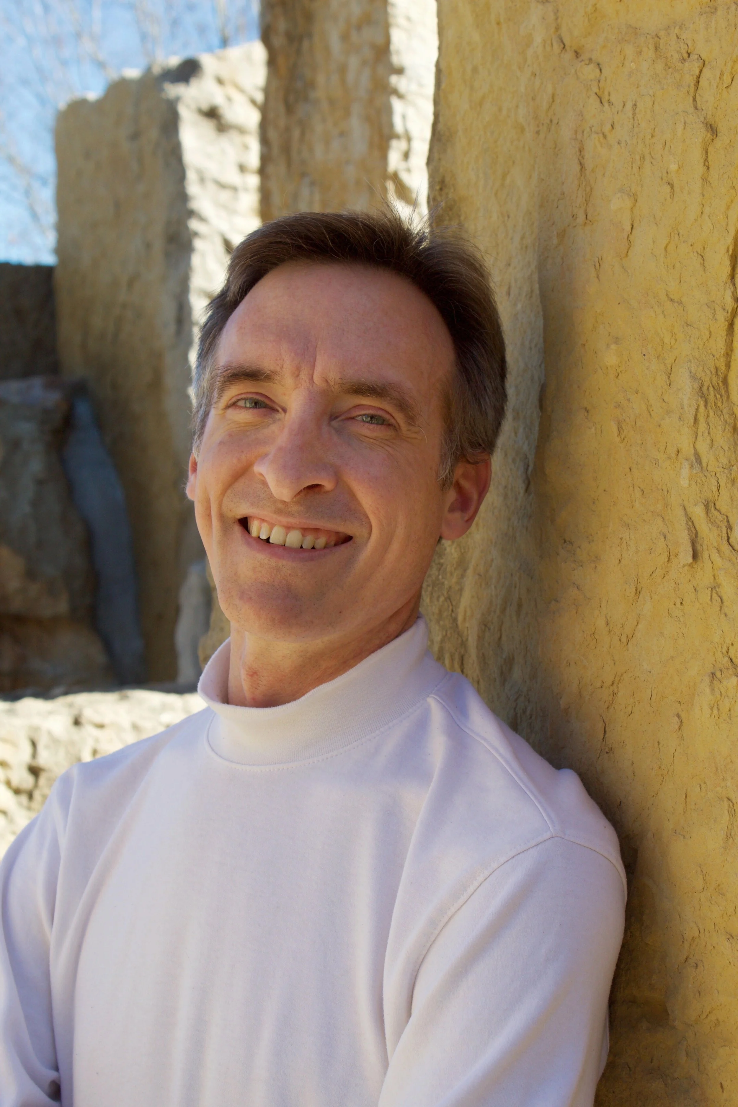 Smiling man with short dark hair, wearing a white turtleneck, leaning against a yellow stone wall outdoors on a sunny day.