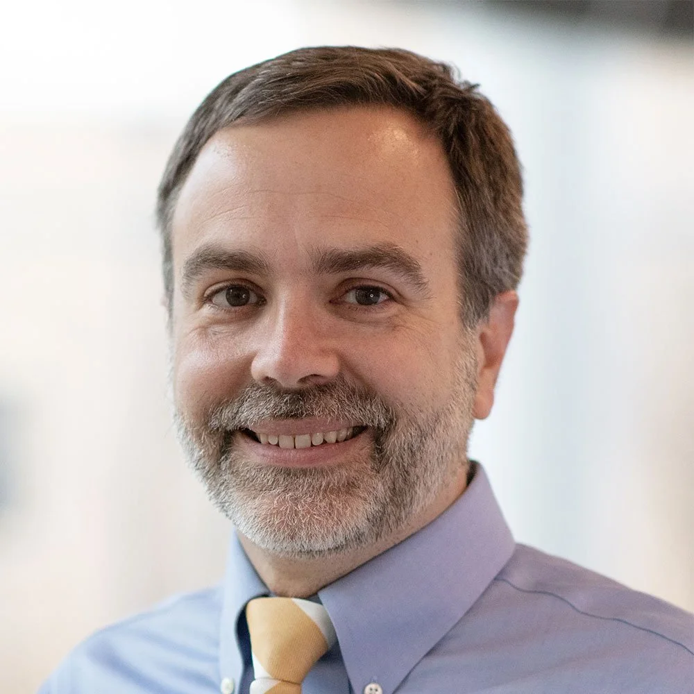 A smiling man with brown hair, a beard, and mustache, wearing a light blue shirt and a yellow tie, in a professional setting.