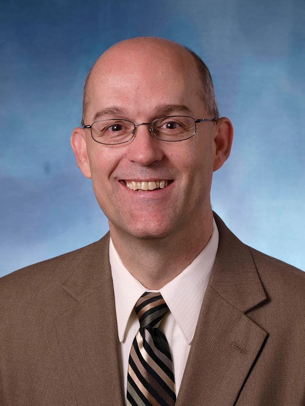 A professional headshot of a smiling man with glasses, wearing a brown suit, white shirt, and striped tie, against a blue gradient background.