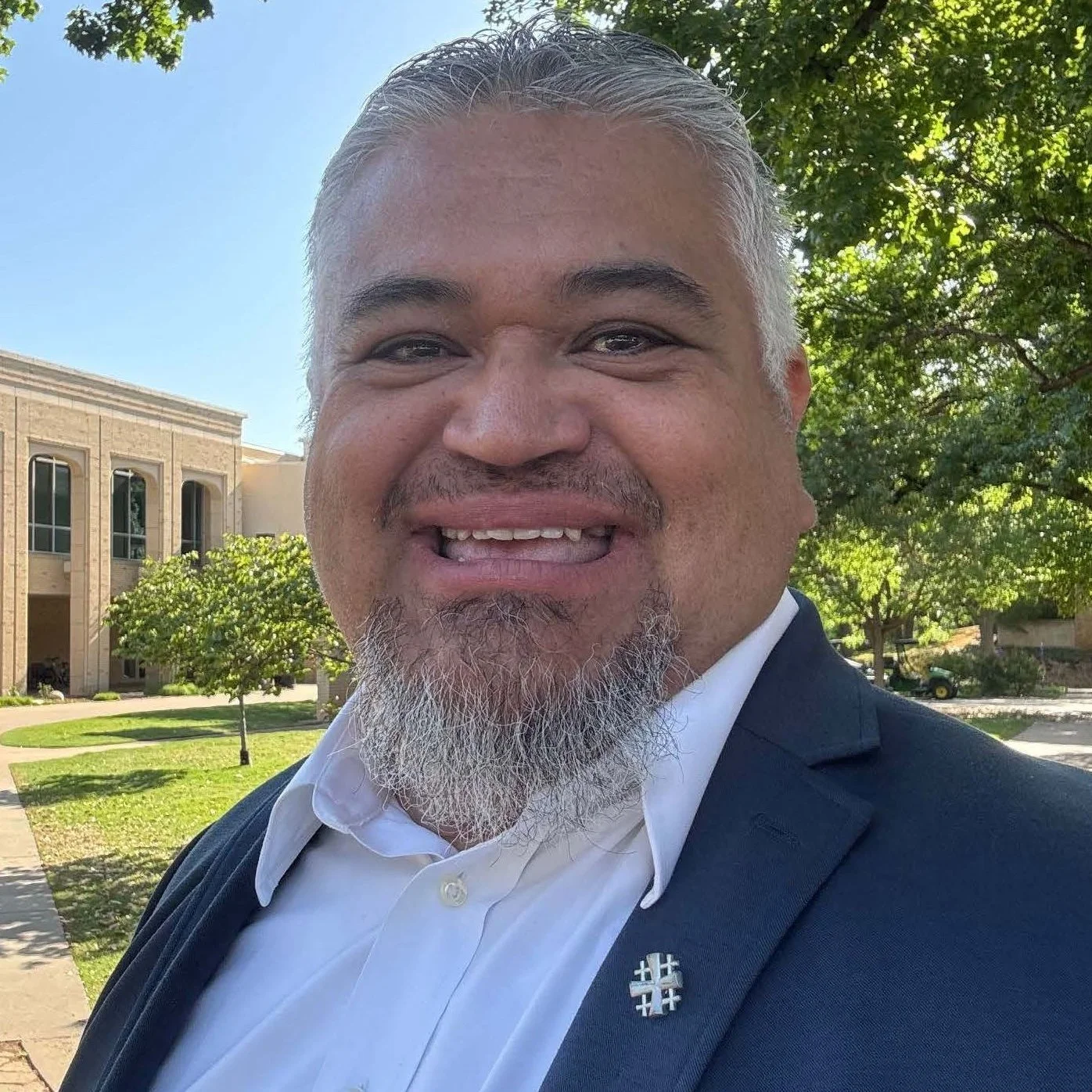 A smiling man with a beard and mustache, gray hair, dressed in a white shirt and dark blazer, standing outdoors with trees, a building, and a golf cart in the background on a sunny day.