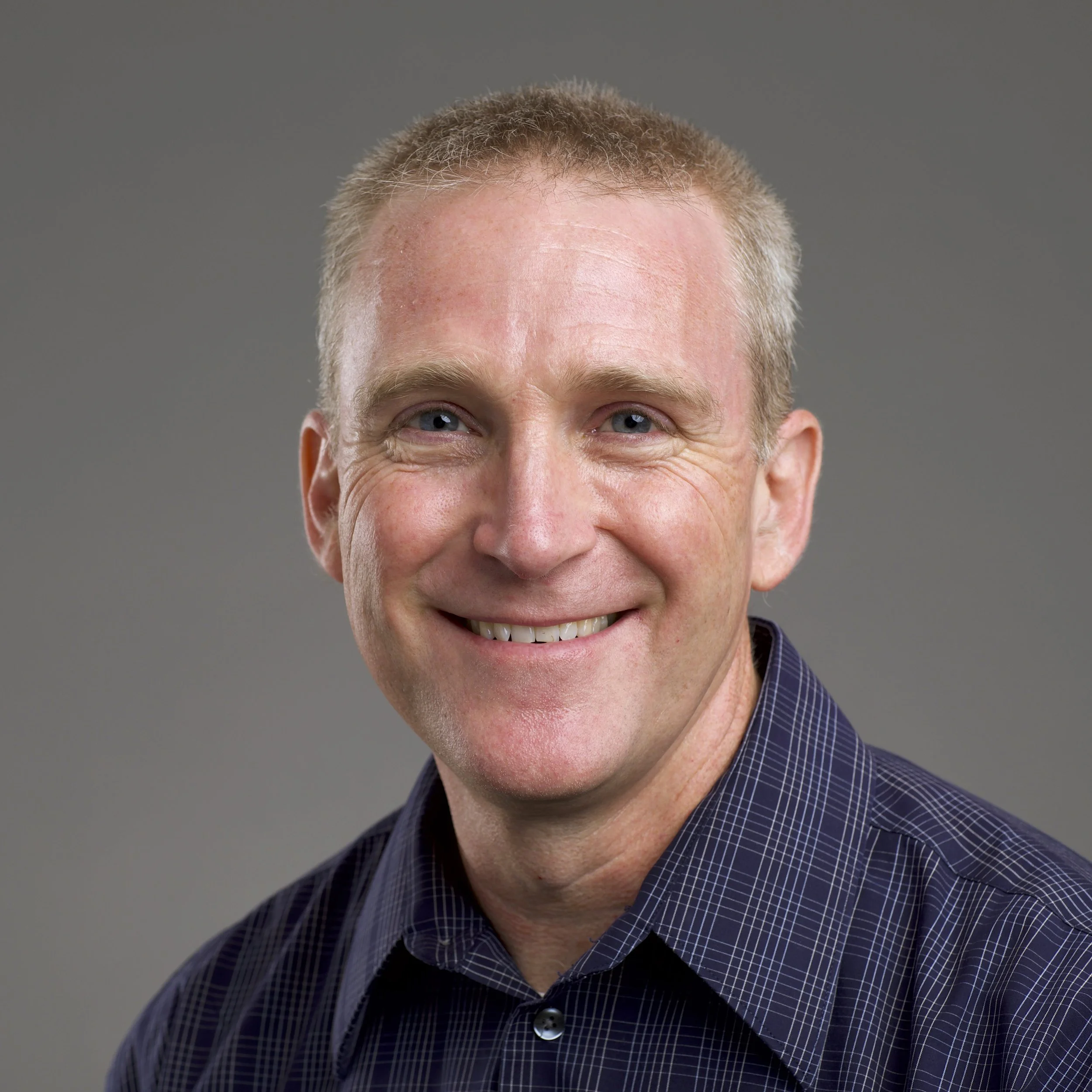 Close-up of a man with short light-colored hair, blue eyes, smiling, wearing a dark checkered shirt, against a plain gray background.