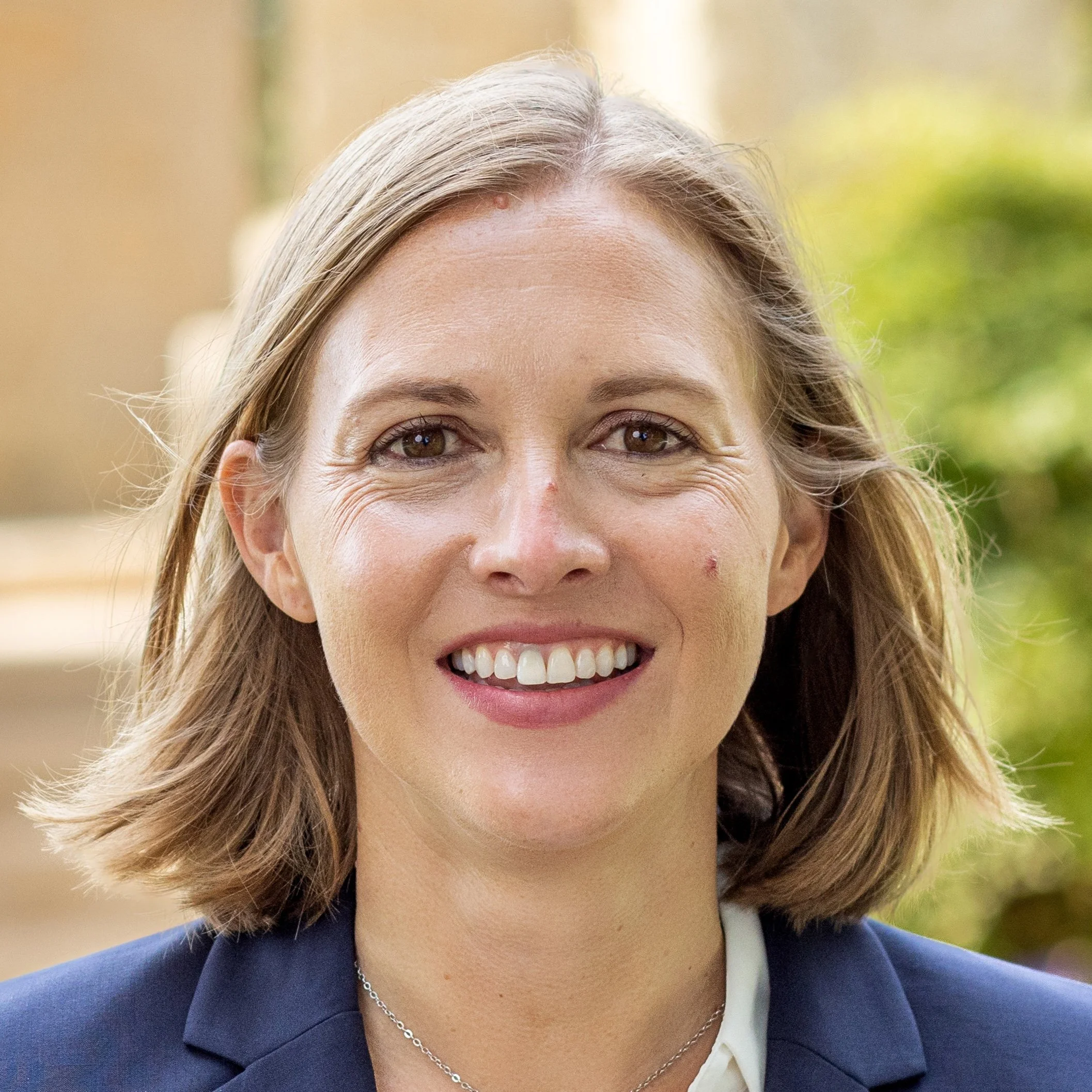 Close-up of smiling woman with brown hair, wearing a navy blazer, white shirt, and silver necklace, outdoors with blurred green and beige background.