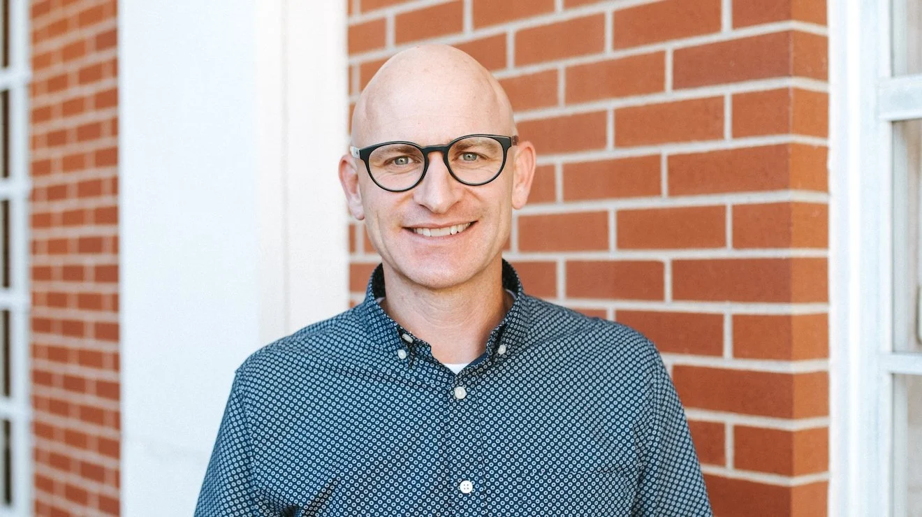 A smiling man with glasses and a blue patterned button-up shirt standing outside in front of a red brick building.