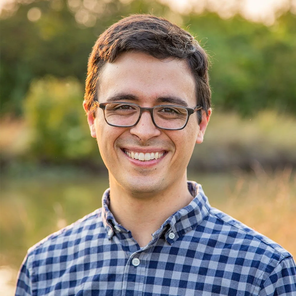 A young man with glasses and dark hair smiling outdoors during golden hour, wearing a checkered shirt.